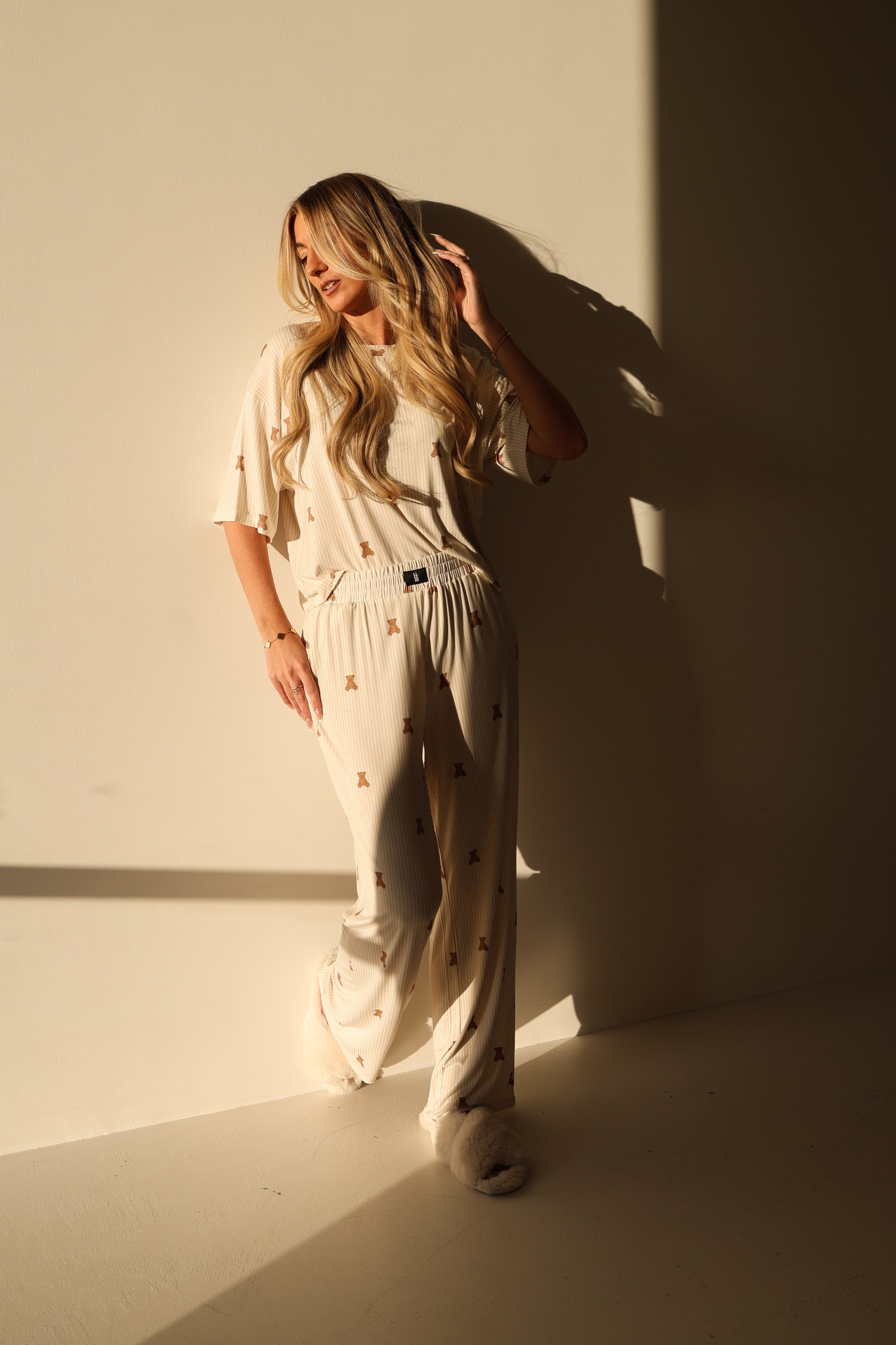 A woman with long blonde hair leans against a sunlit wall, wearing the forever french baby Women's Boxy Tee Lounge Set | Teddy with small patterns and fluffy slippers, as soft shadows are cast behind her.