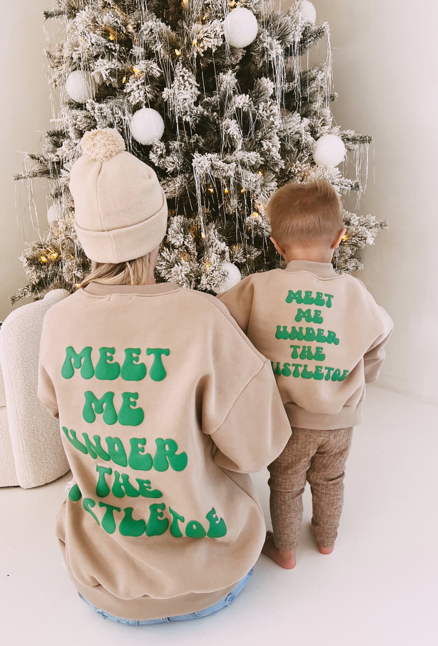 An adult and a child, both donning festive "Meet Me Under The Mistletoe" sweatshirts by forever french baby, stand with their backs to the camera. They gaze at a Christmas tree decorated with white ornaments and tinsel.