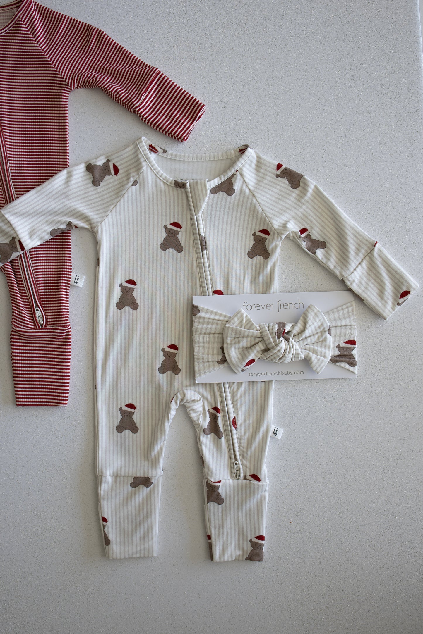 A cream baby onesie with a brown teddy bear print and the forever french baby Bamboo Head Wrap | Santa Teddy is displayed next to a red and white striped baby outfit on a light surface, perfect for celebrating your little one's first Santa Teddy moments.