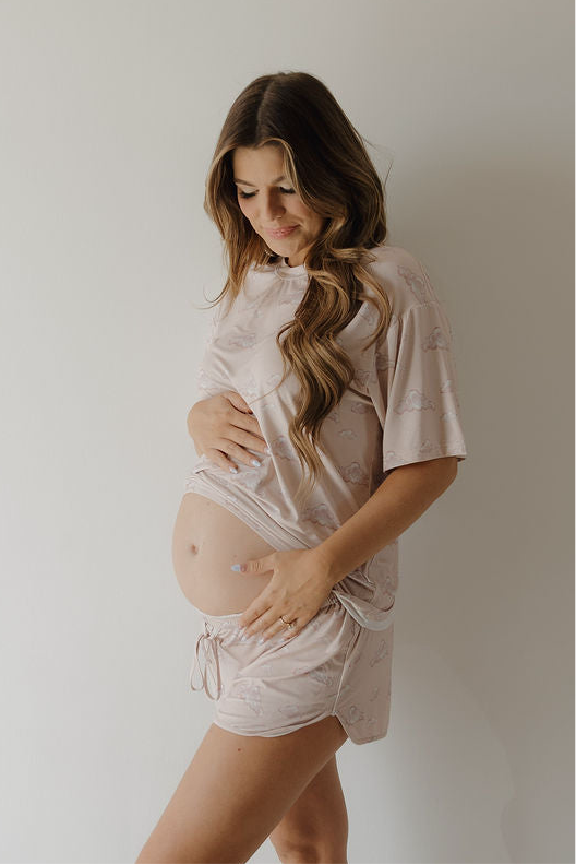 A pregnant woman with long brown hair stands sideways, smiling softly and cradling her baby bump. She wears the Kendy 3 Women's Bamboo Short Set in Ruby—loose, breathable light pink pajamas with short sleeves and shorts—against a plain white background.