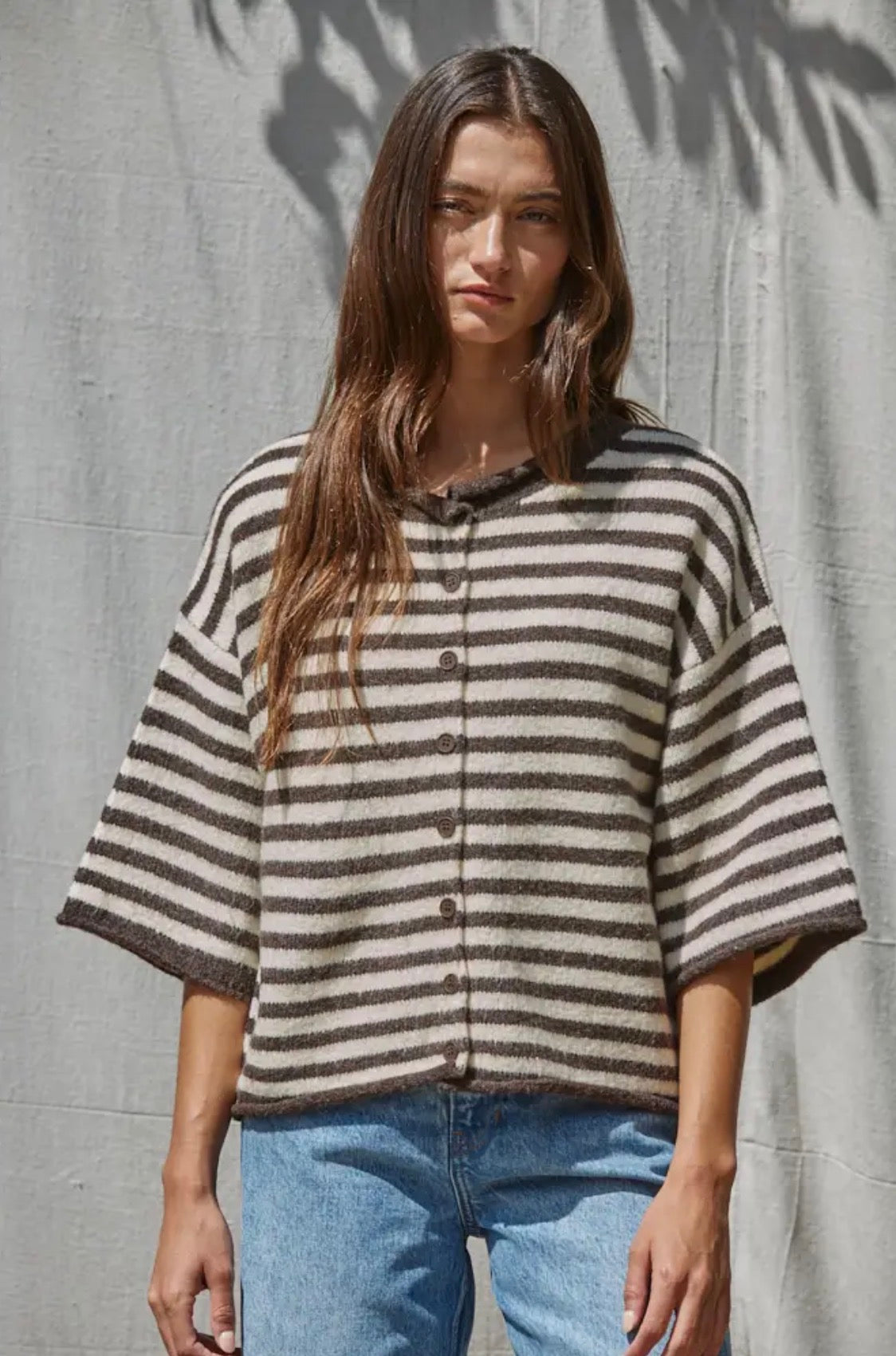 A woman with long brown hair wears the by Together Knit Sweater Striped Quarter Sleeve Cardigan—featuring loose, wide sleeves and brown and beige stripes—paired with light blue jeans, standing in front of a light gray fabric backdrop.