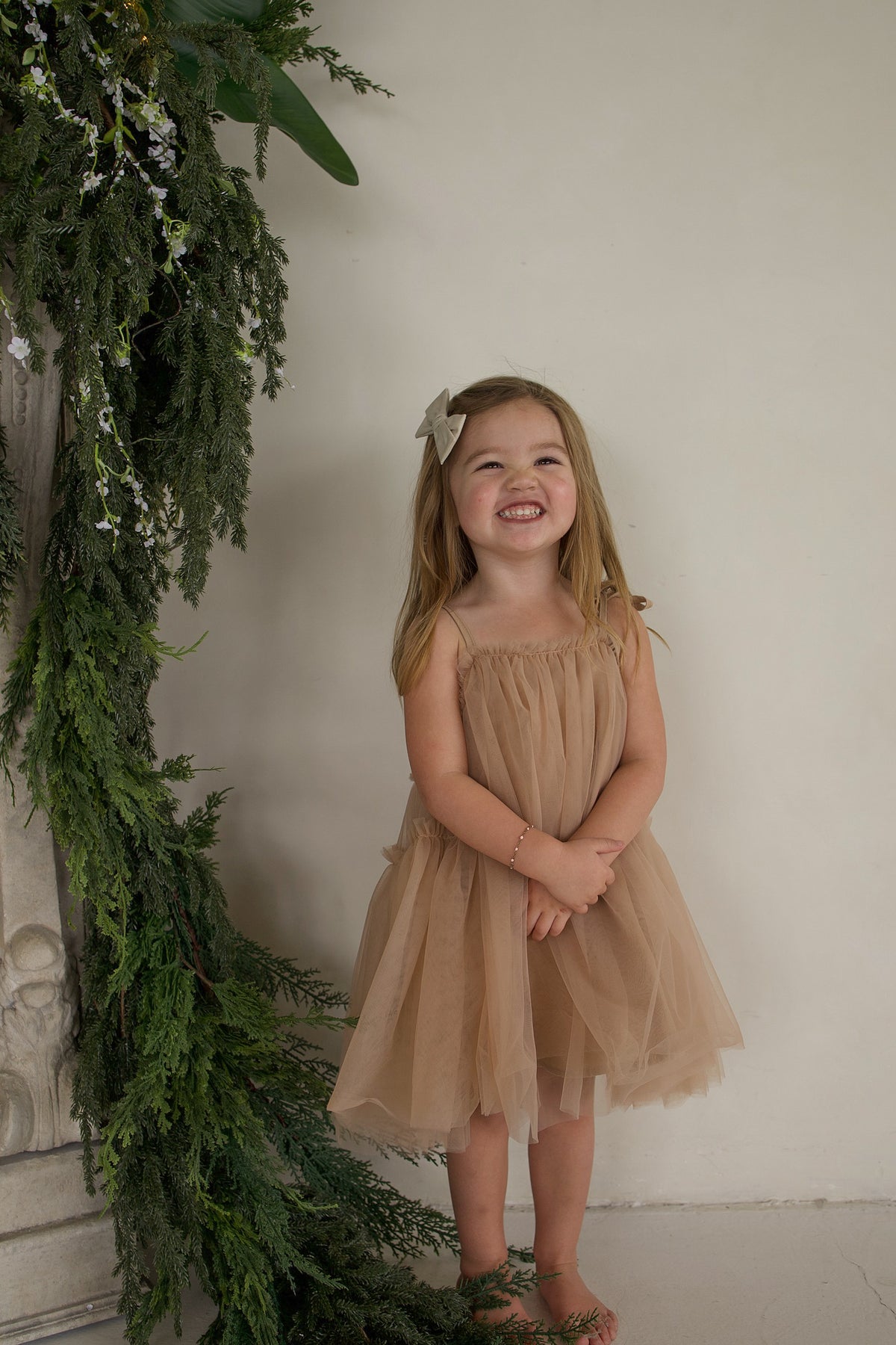 A young girl smiles while wearing a tan sleeveless Tutu Dress | Teddy with a matching bow in her hair.