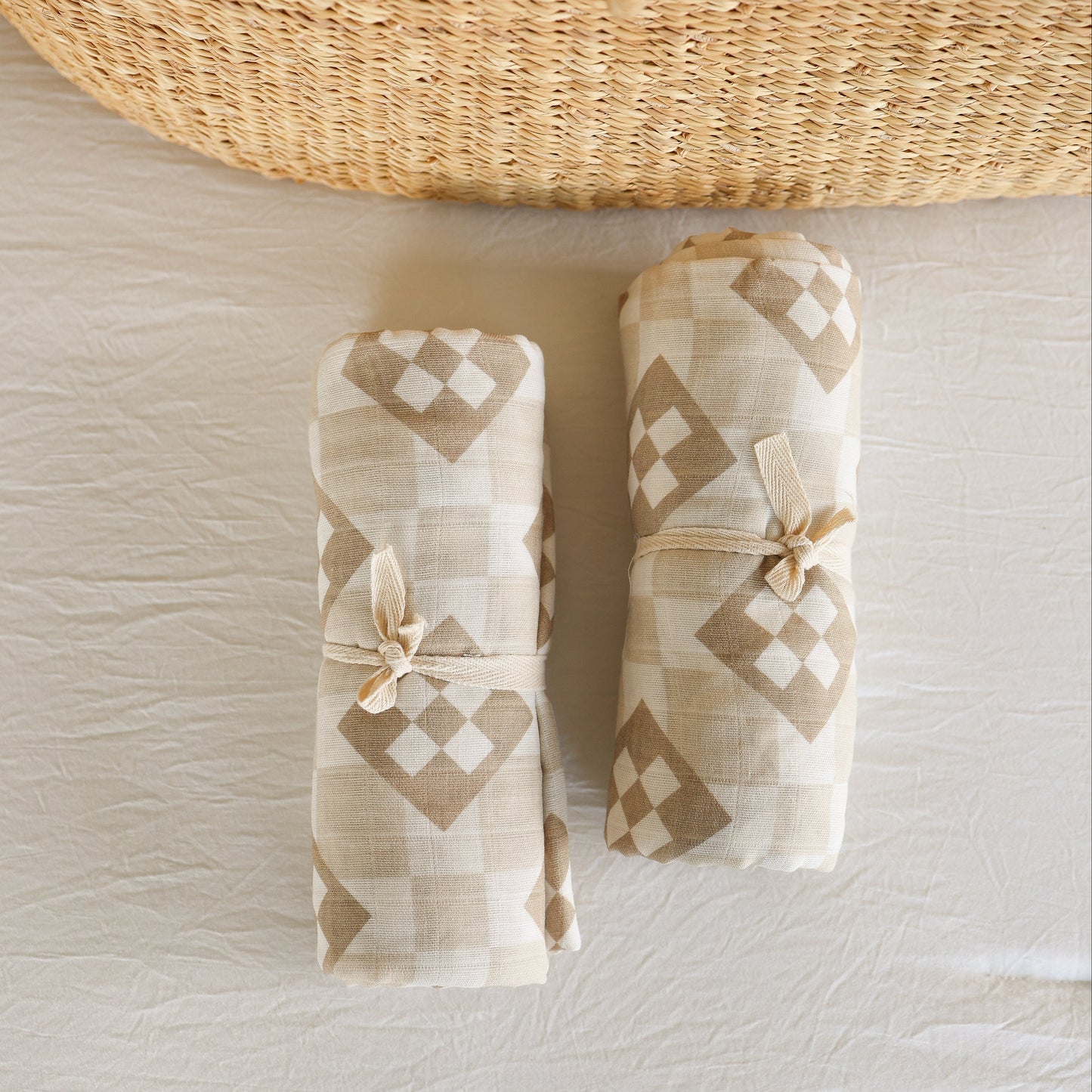 Two rolled forever french baby Muslin Swaddle | Cottage Quilt blankets, featuring a beige and white diamond pattern and tied with fabric bows, rest on a light textured surface beside a woven basket.