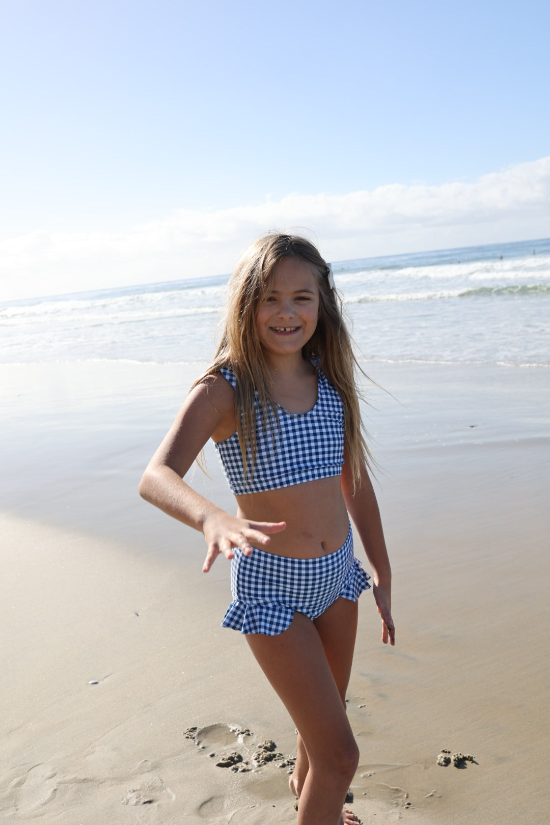 A girl smiles on a sandy beach wearing the forever french baby Girls Two Piece Swimsuit in Blue Gingham, with gentle waves and a blue sky in the background.