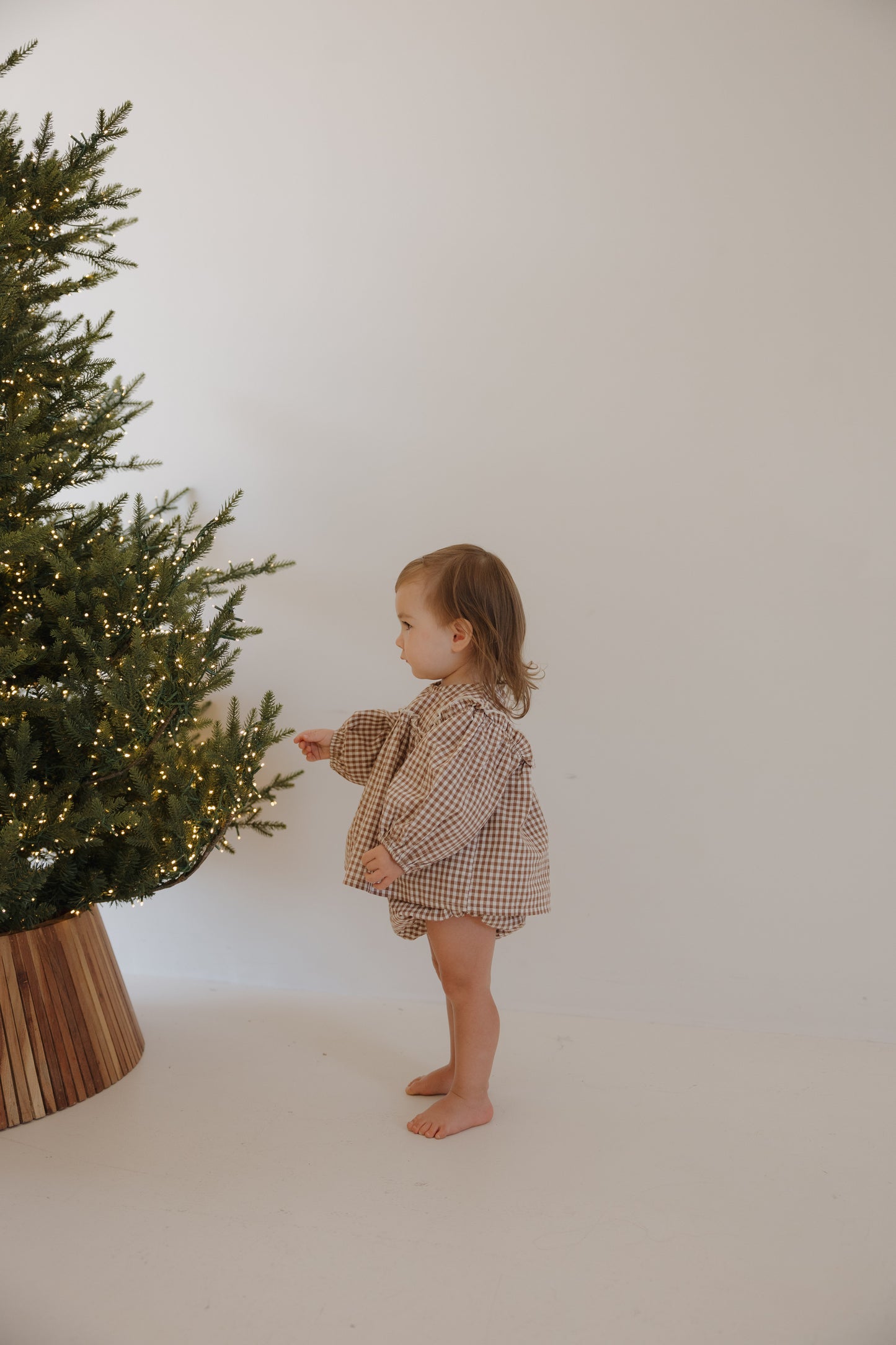 A barefoot toddler wears the forever french baby Bloomer Set in Honey Gingham, reaching for the branches of a Christmas tree adorned with small white lights against a plain white background.