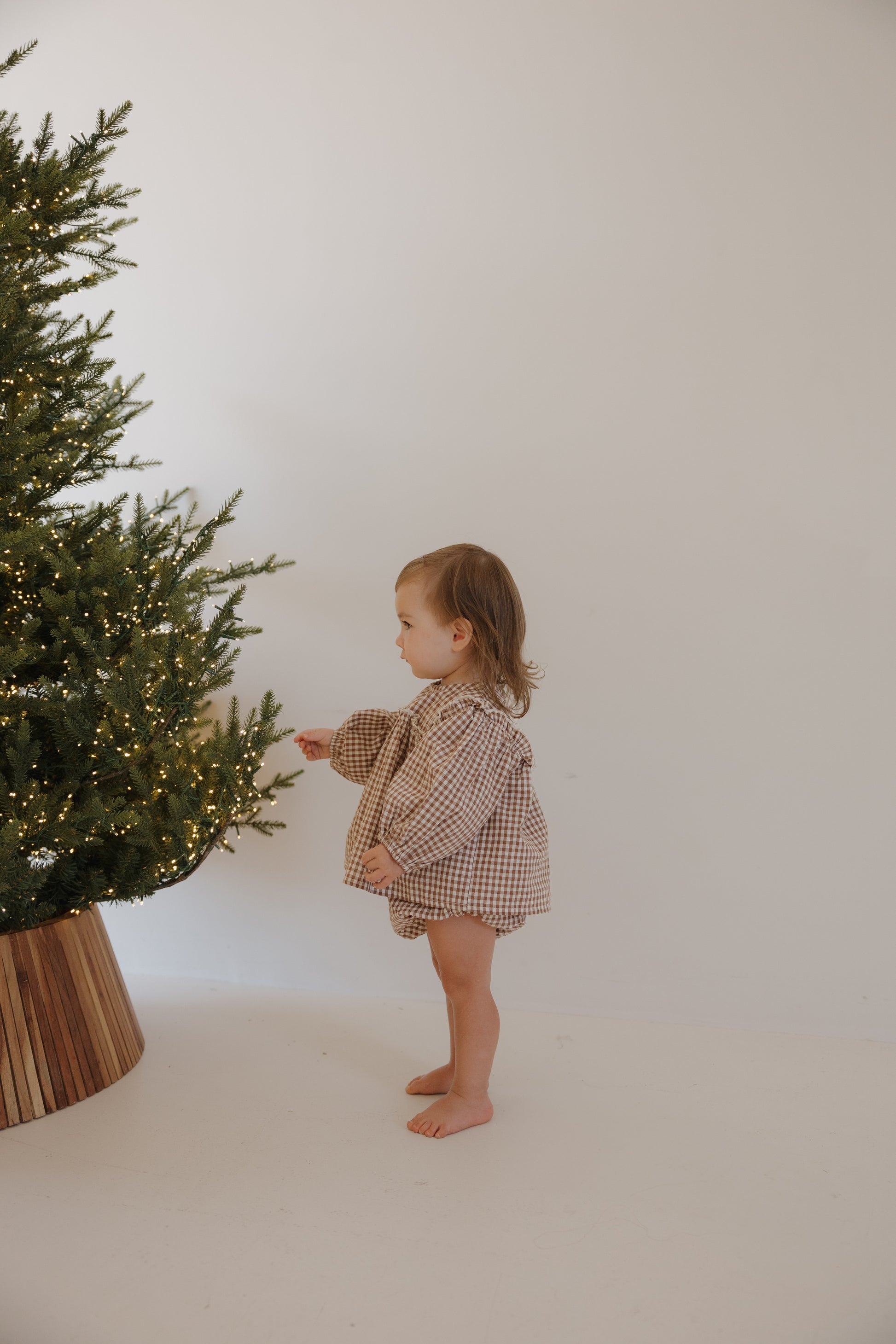 A barefoot toddler wears the forever french baby Bloomer Set in Honey Gingham, reaching for the branches of a Christmas tree adorned with small white lights against a plain white background.