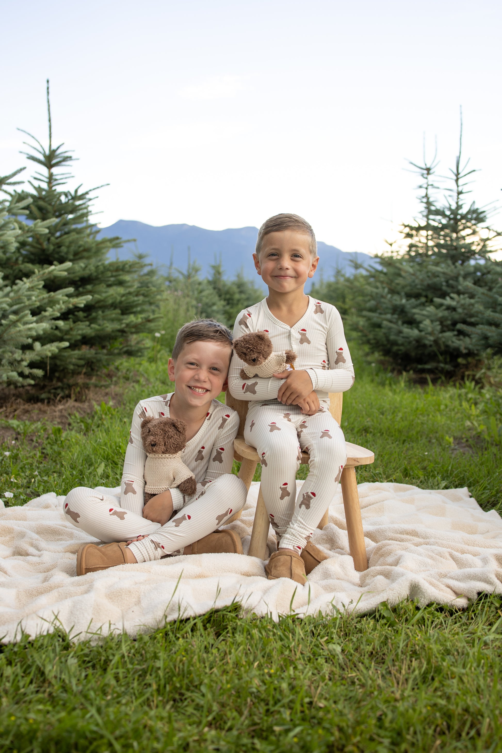 Two young boys smile at the camera among small pine trees and mountains, wearing matching forever french baby Bamboo Two Piece Pajamas | Santa Teddy. One sits on a stool, the other on a blanket; both hold Santa Teddy bears as they enjoy the outdoor scene.