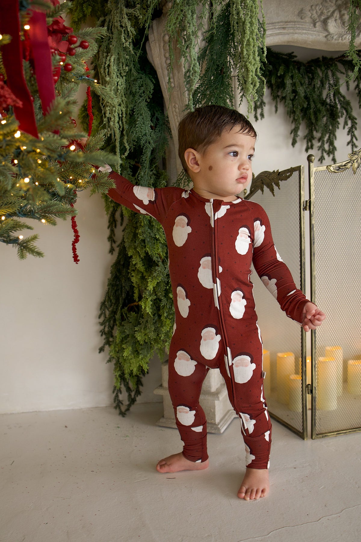 A toddler wearing a long-sleeved, dark red Bamboo Zip Pajamas | Vintage Santa romper patterned with vintage Santa Claus illustrations.