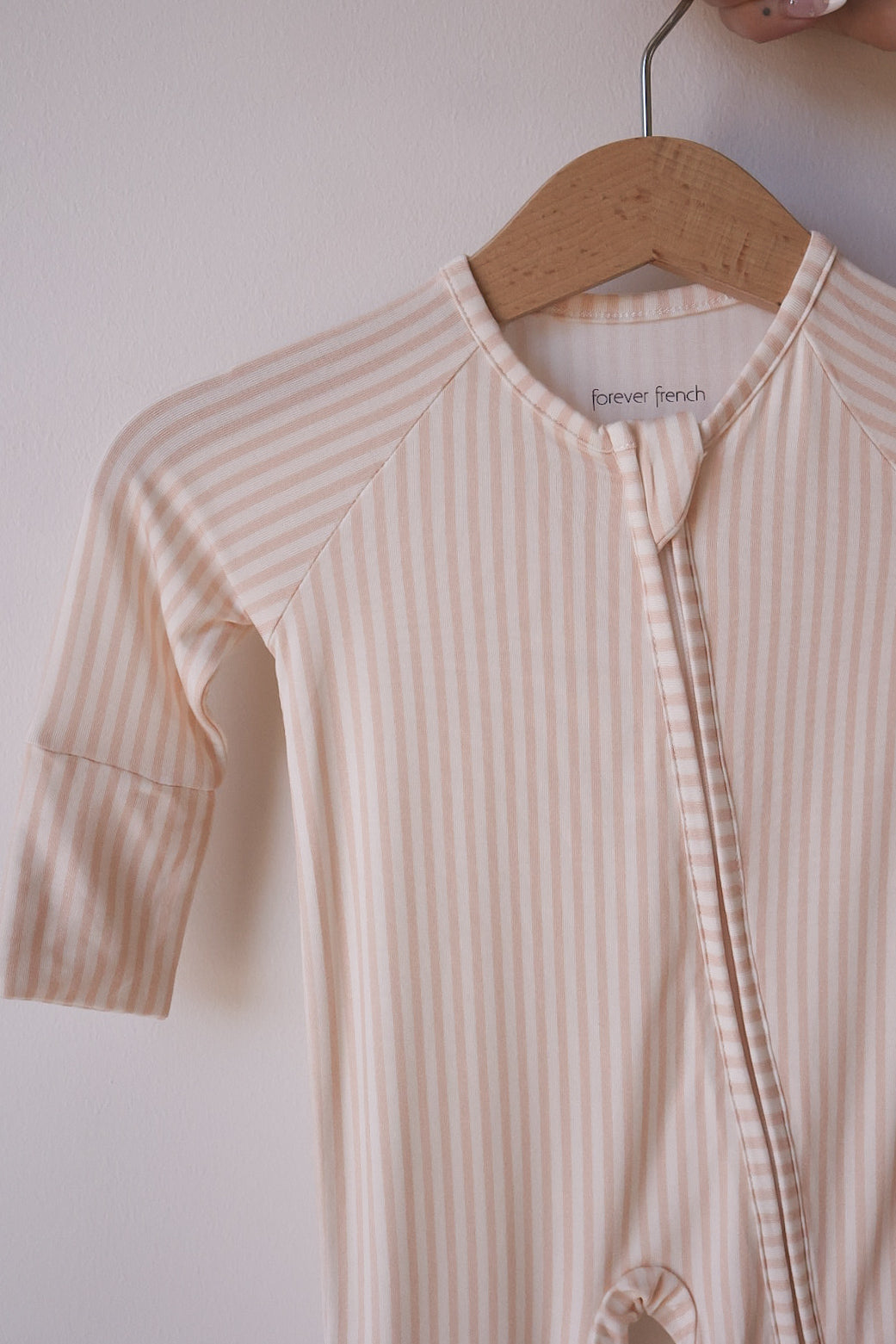 The Bamboo Zip Pajamas | Sadie Stripe by forever french baby, featuring beige and white stripes and long sleeves, hangs on a wooden hanger against a light wall.