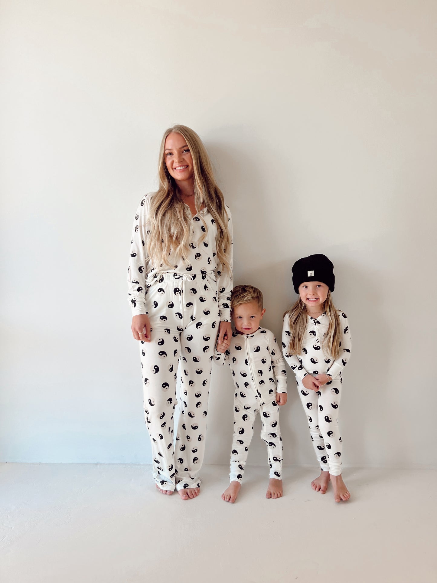 A woman and two children stand against a white wall, all wearing matching Women's Bamboo Pajamas | Black & White Yin Yang from forever French baby. One child is wearing a black beanie, and all are smiling. The floor is also white, enhancing the minimalistic background focused on the trio in coordinated outfits with a black and white design.