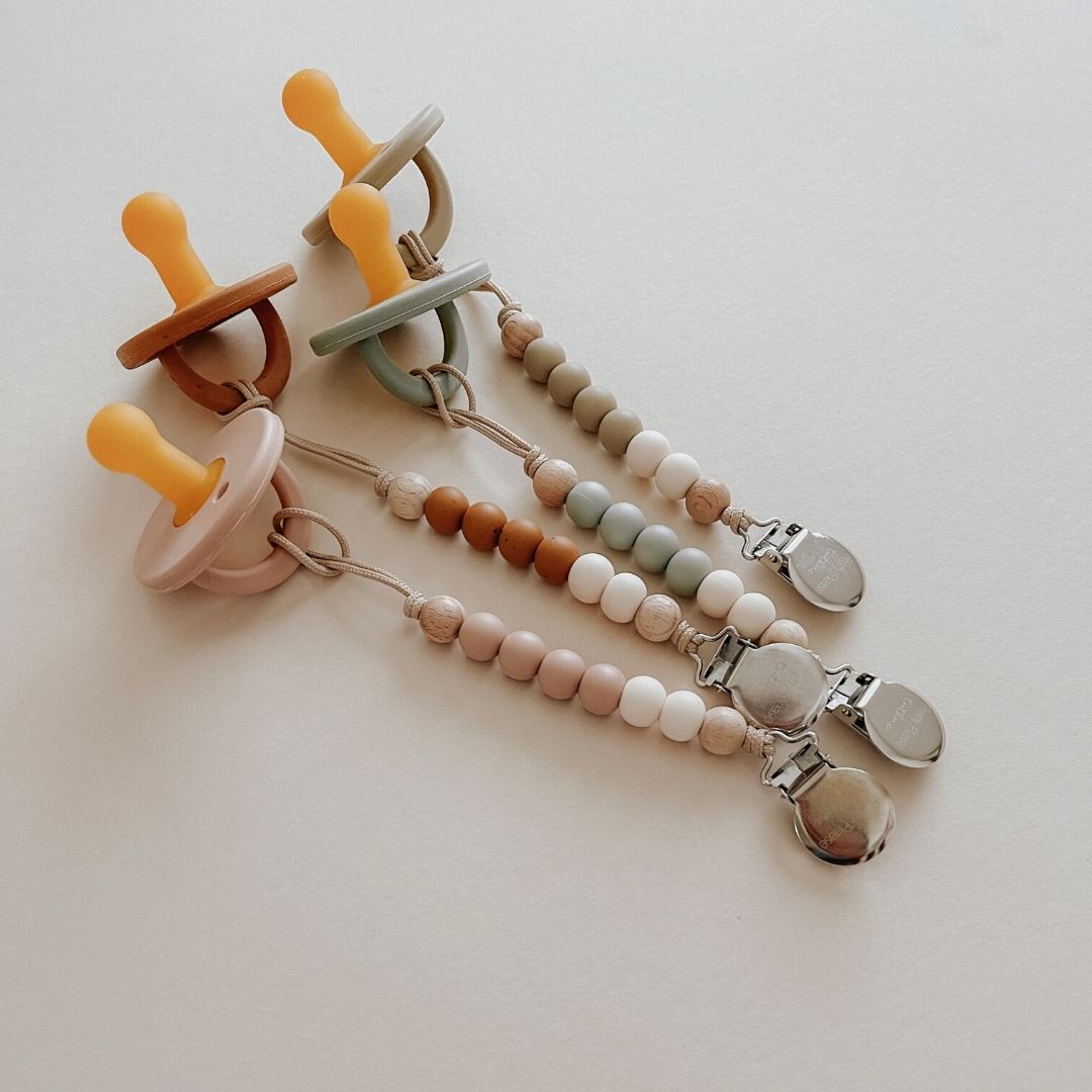 Image of three Modern Pacifier | Speckled Almond by forever french baby. Each silicone pacifier is attached to a beaded clip chain, featuring beads in soft pastel colors including beige, mint green, and peach. The metallic clips at the end are designed to attach securely to a baby's clothing, making them essential baby essentials.