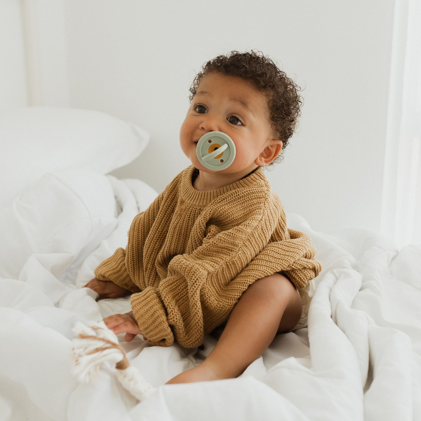 A baby with curly hair, dressed in a cozy beige sweater, sits on a white, quilted bed. The baby holds the Modern Pacifier | Fawn by forever french baby in their mouth and looks inquisitively towards the camera. The background is minimal and bright, creating a soft, warm atmosphere—perfect for showcasing baby essentials.