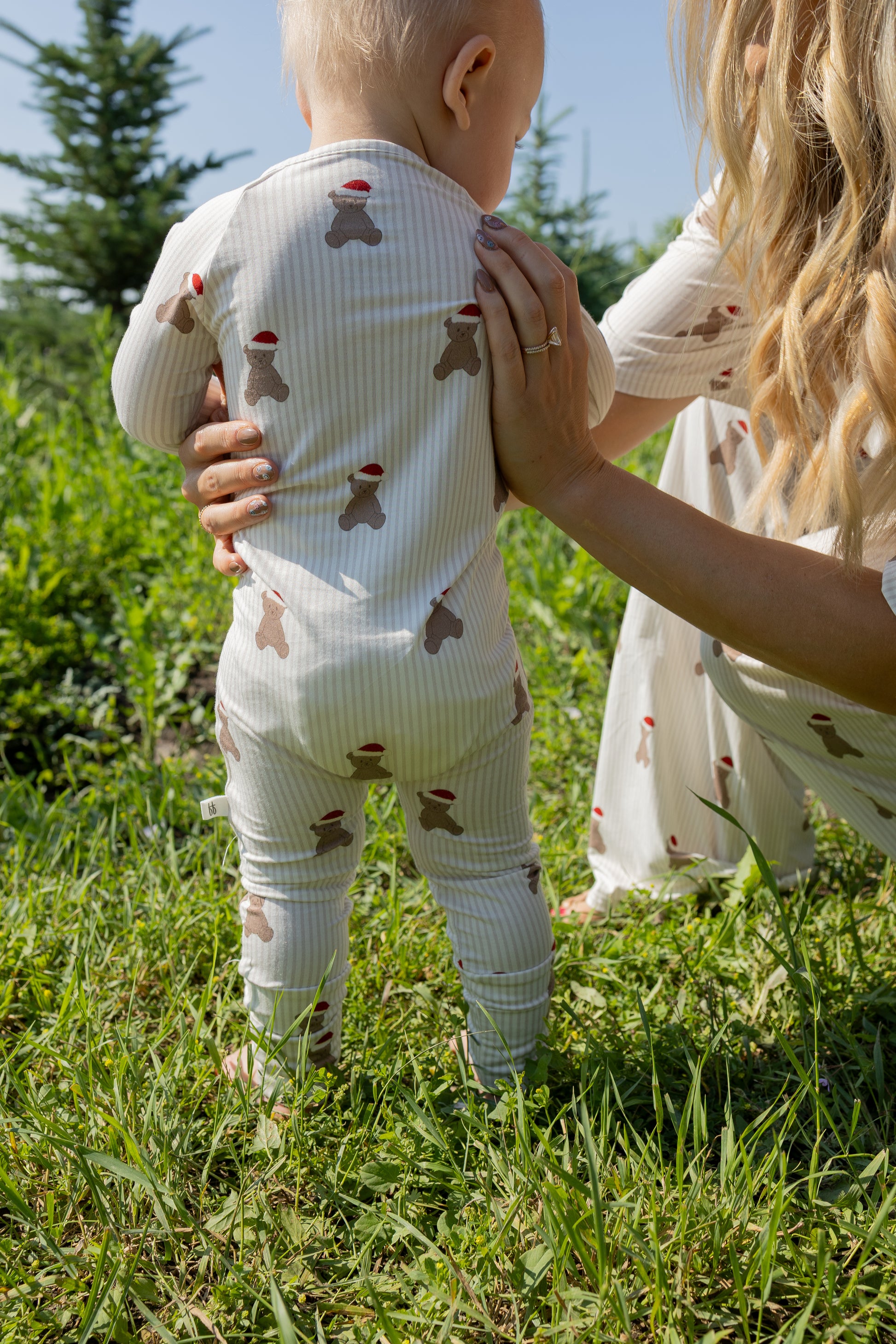 A baby dressed in forever french baby's Bamboo Zip Pajamas | Santa Teddy stands on grass, supported by an adult with long blonde hair. Both are wearing matching hypoallergenic outfits featuring the Santa Teddy print. Green trees and plants fill the background beneath a clear blue sky.
