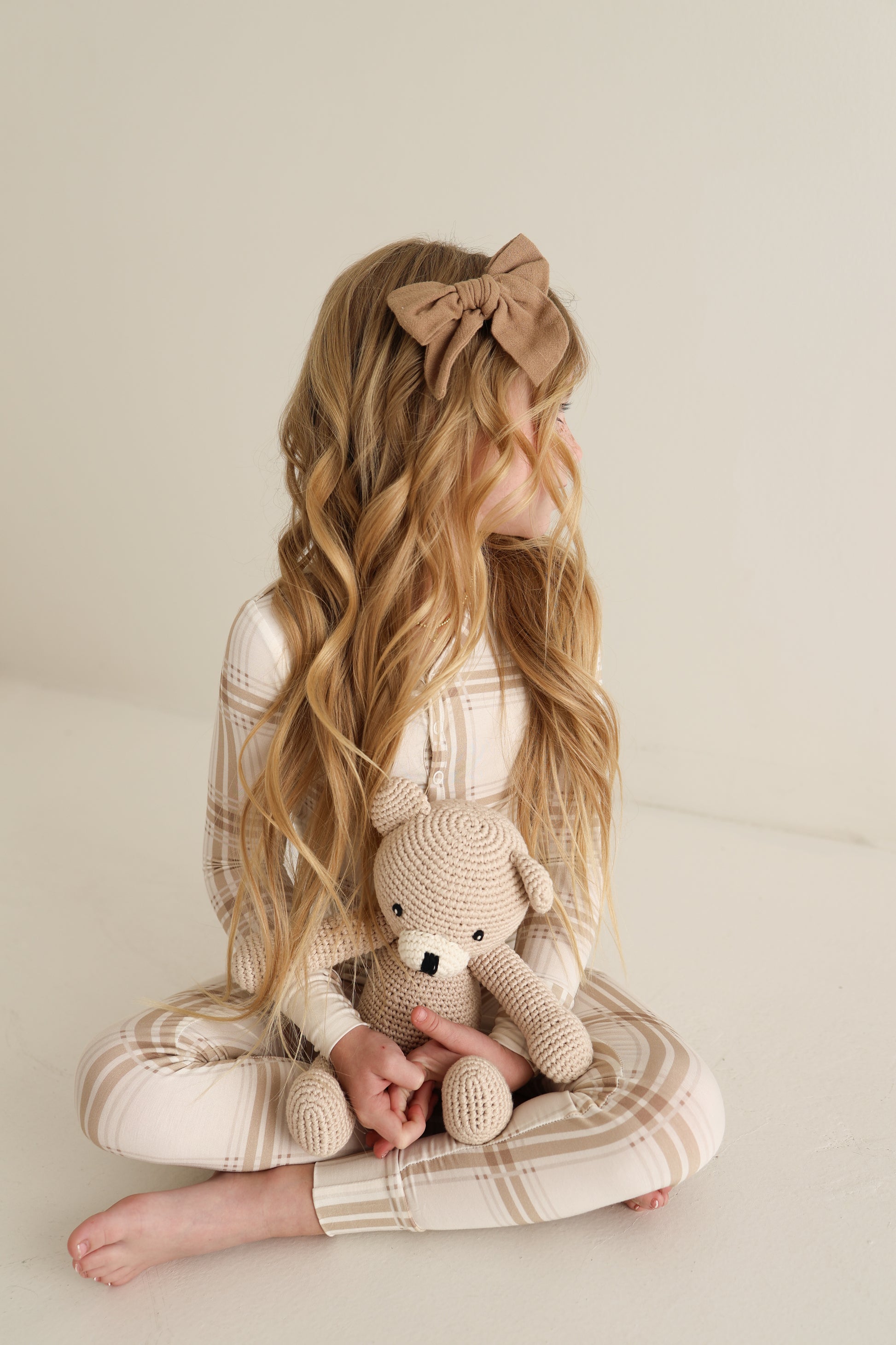A young girl with long wavy blonde hair, wearing a brown bow and forever french baby Bamboo Two Piece Pajamas in Ellis Plaid, sits on the floor holding a knitted teddy bear, looking left against a plain light background.