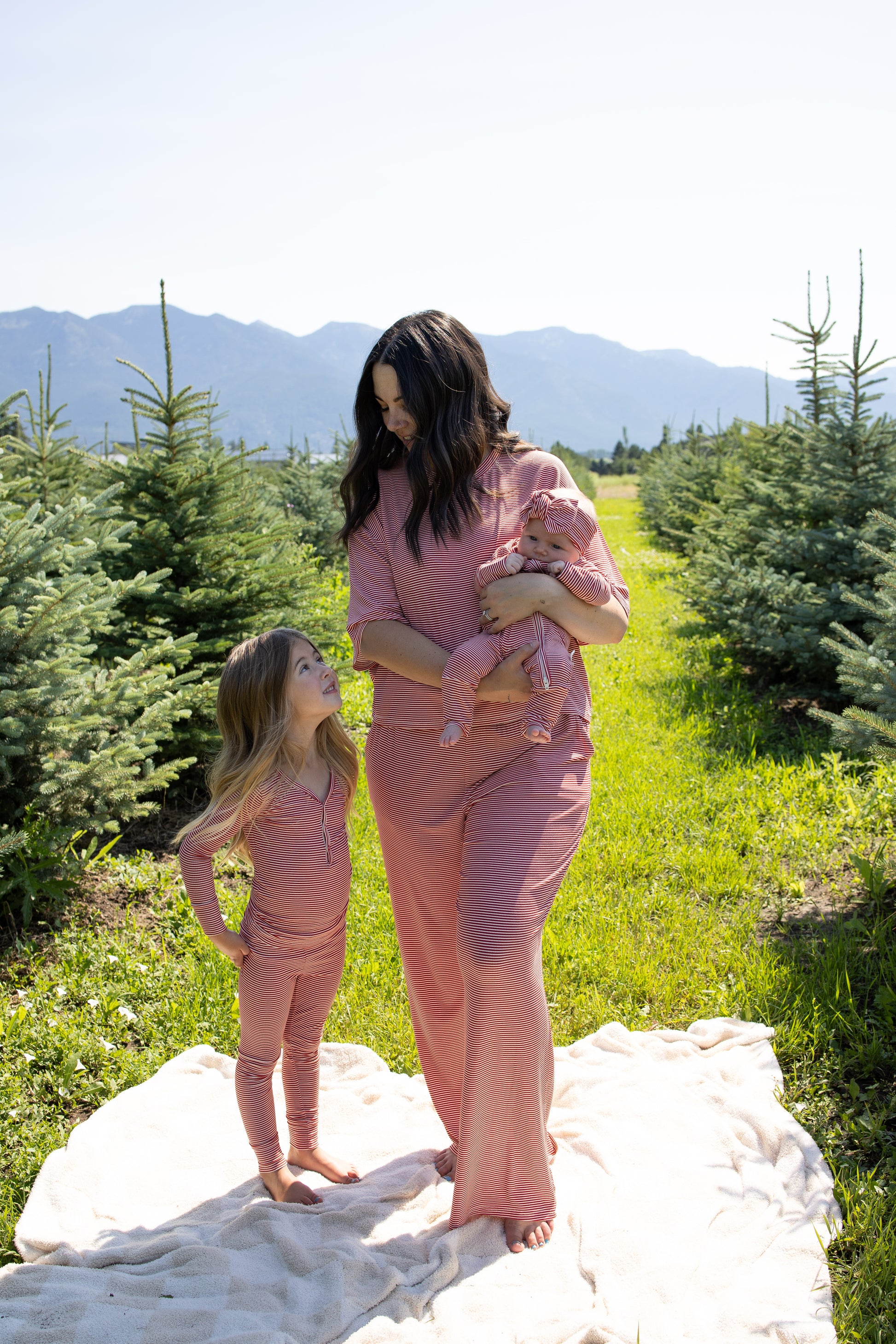 A woman holding a baby stands next to a young girl, all dressed in matching forever french baby Women's Boxy Tee Lounge Sets in Peppermint Stripe Ribbed, posing on a blanket among small evergreen trees with mountains visible in the background.