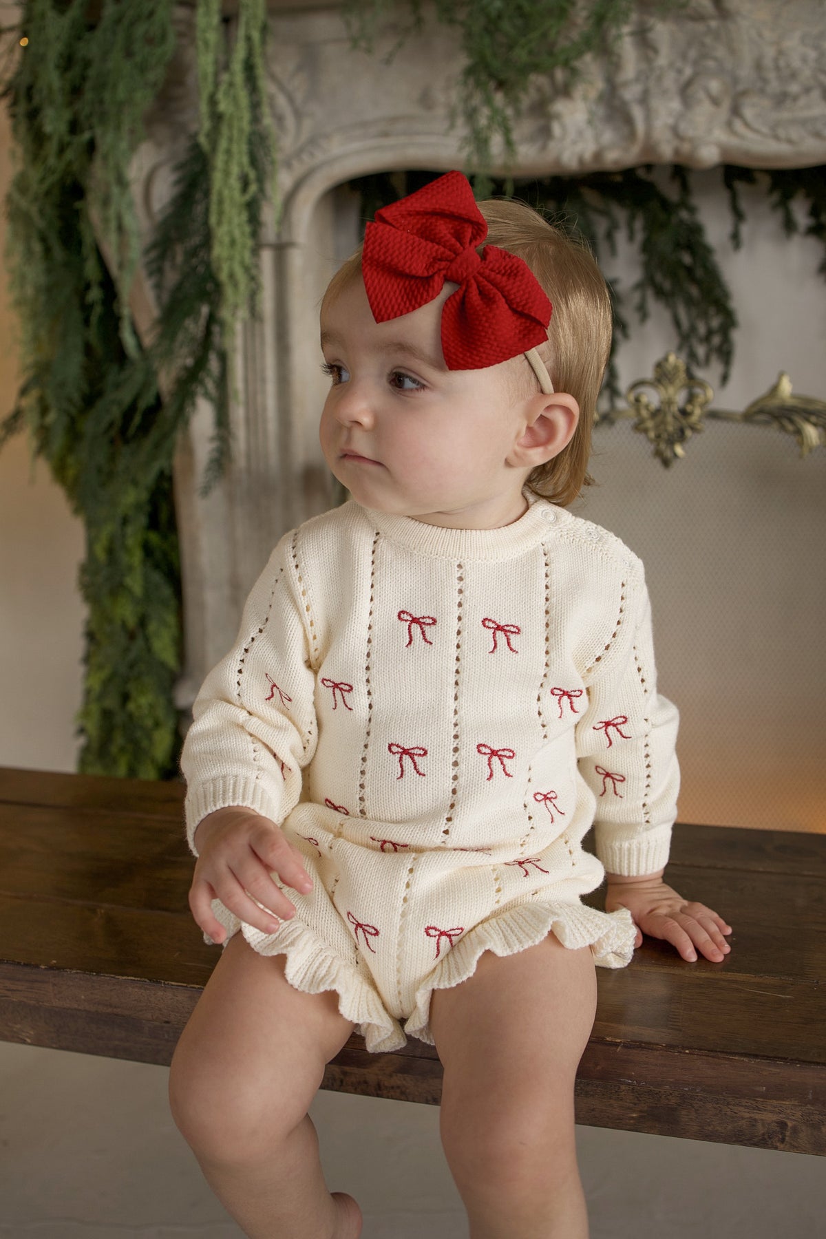 A toddler wearing a Knit Ruffle Romper | Candy Cane sits on a wooden bench in a soft knit baby romper with a red bow pattern and a matching red hair bow.