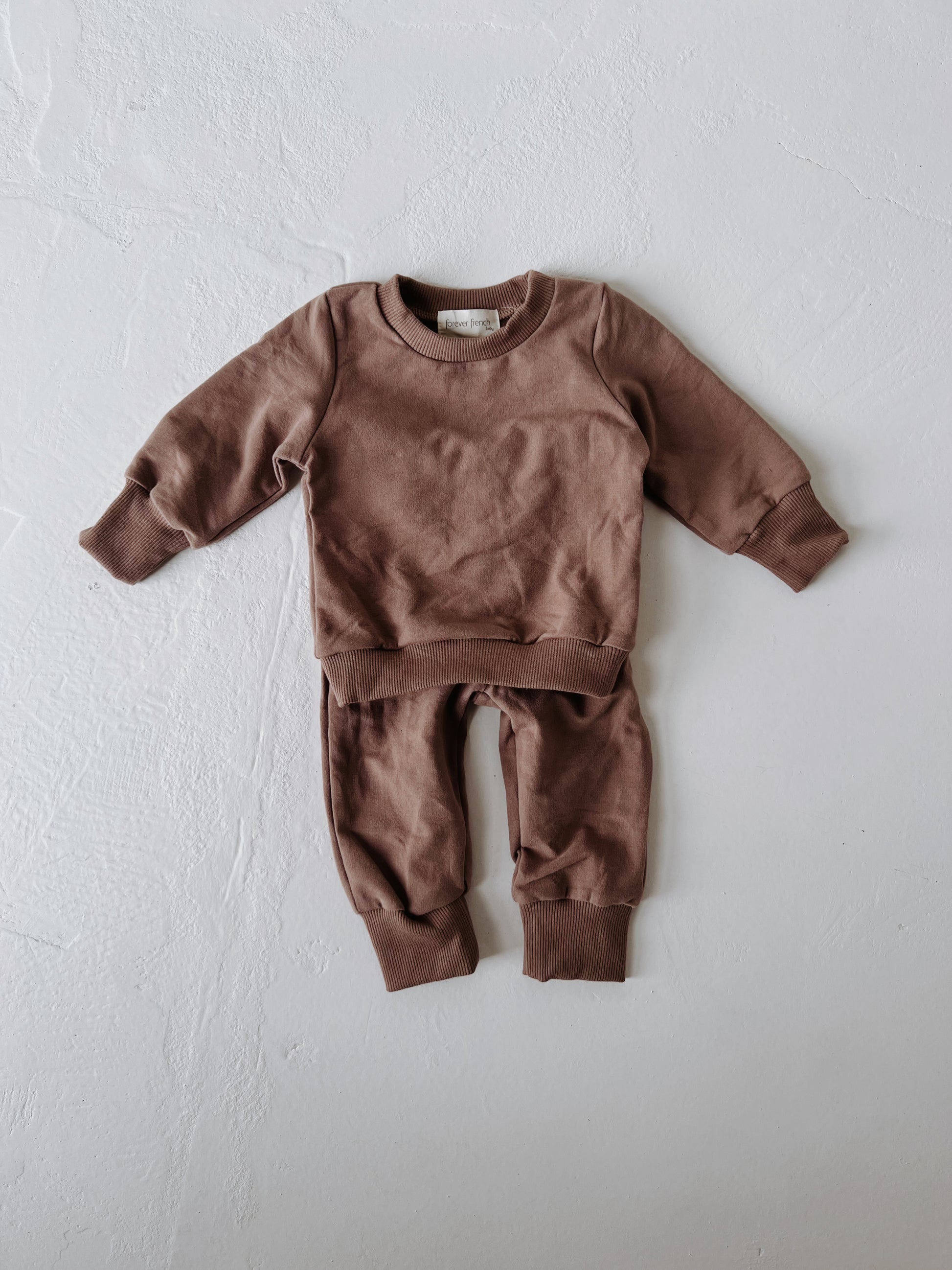 The forever french baby Child Sweat Set | Hot Cocoa is a brown toddler outfit with a long-sleeved sweatshirt and matching pants, shown laid flat on a textured white surface—ideal for coordinated family style.
