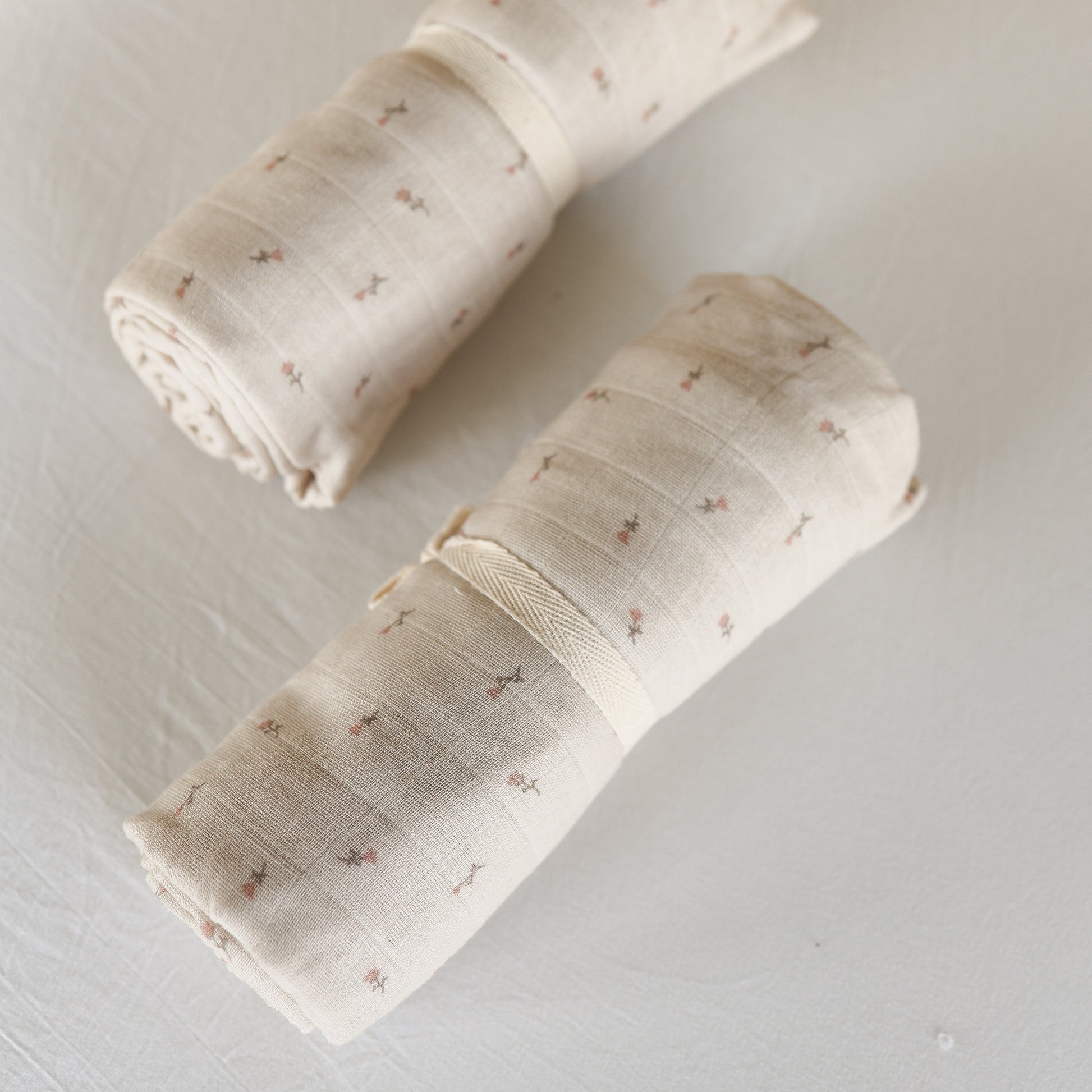 Two “Muslin Swaddle | Lucy” blankets by forever french baby, featuring a beige hue with grid and floral patterns, are rolled up and held with cream bands, displayed on a white textured background.