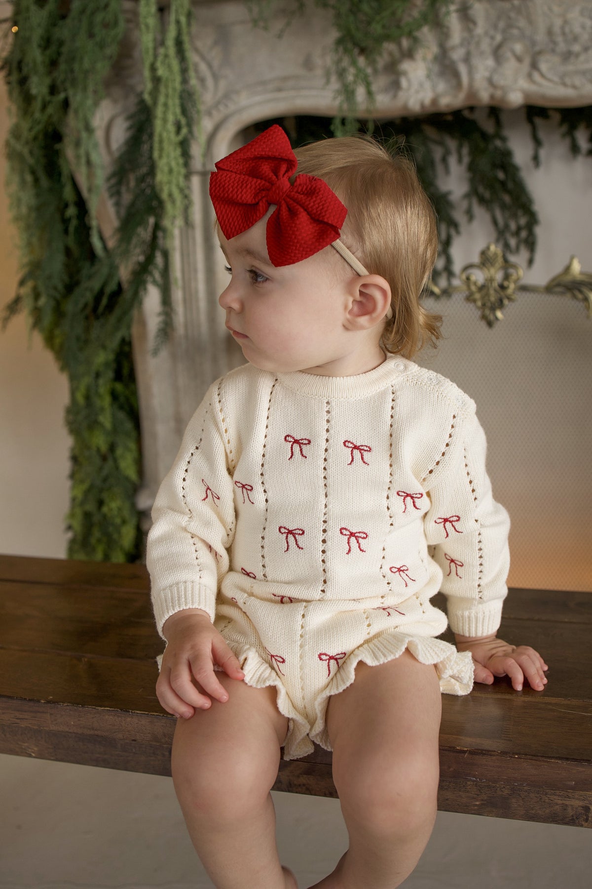 A toddler wearing a cream-colored Knit Ruffle Romper | Candy Cane with red bow patterns and a large red bow headband.