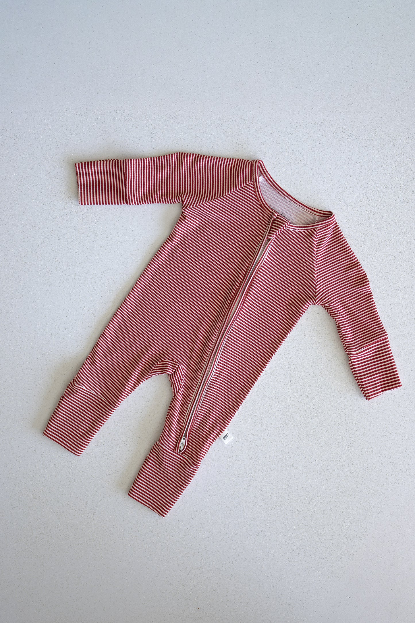 The forever french baby Bamboo Zip Pajamas in Peppermint Stripe Ribbed feature red and white stripes, long sleeves and legs, and are crafted from ultra-comfortable ribbed bamboo fabric. The design includes a diagonal zipper running from the neck to the left leg, shown laid flat on a light background.