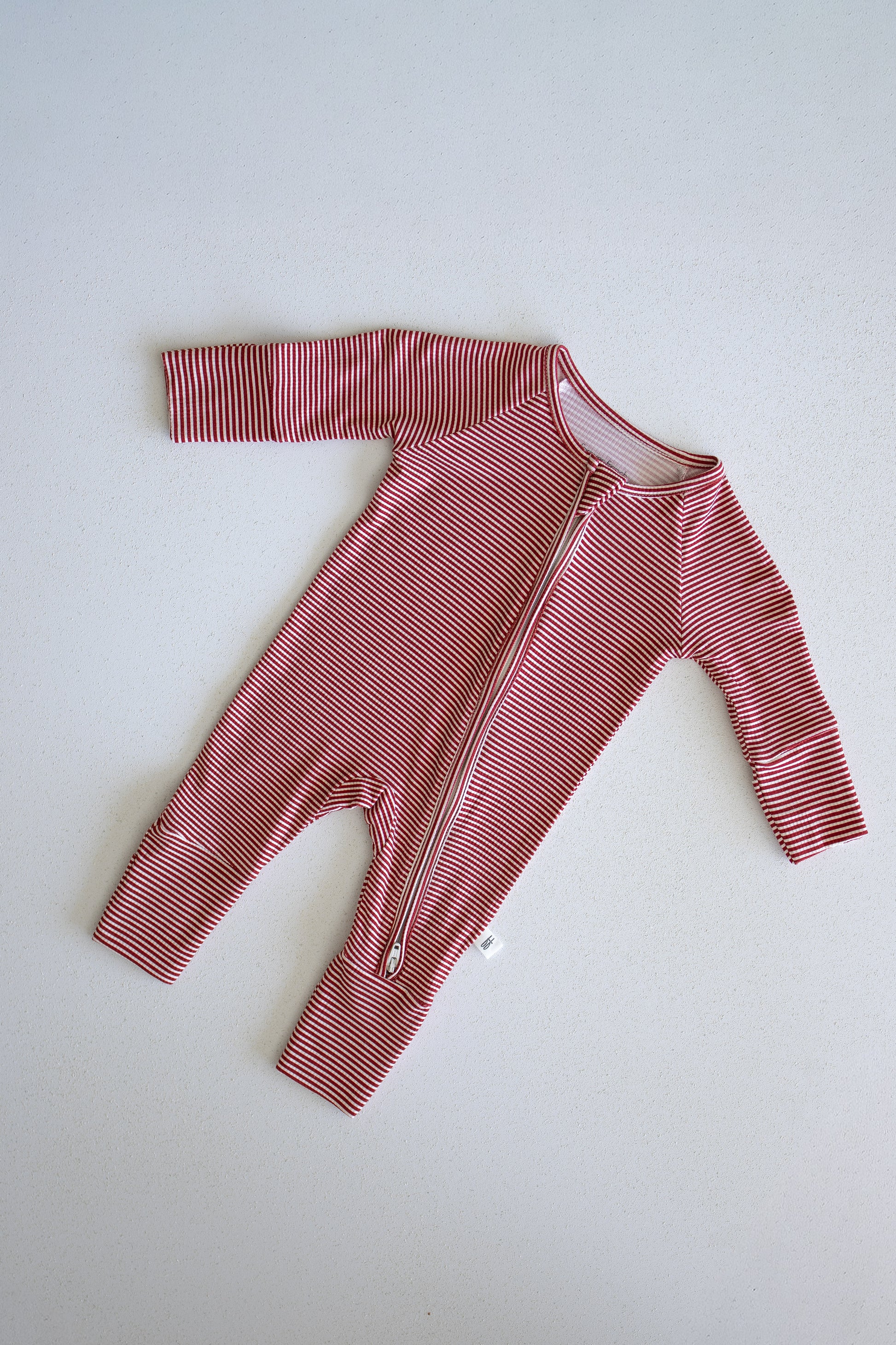 The forever french baby Bamboo Zip Pajamas in Peppermint Stripe Ribbed feature red and white stripes, long sleeves and legs, and are crafted from ultra-comfortable ribbed bamboo fabric. The design includes a diagonal zipper running from the neck to the left leg, shown laid flat on a light background.