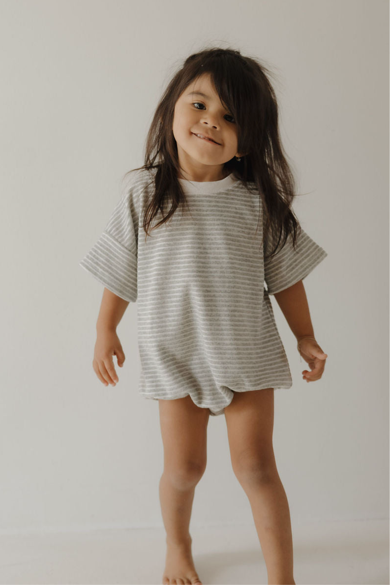 A young child with long dark hair stands barefoot against a plain backdrop, wearing the forever french baby Terry T-Shirt Romper | Grey Stripe—loose-fitting, short-sleeved, stylish, and ideal for family moments and easy diaper changes.