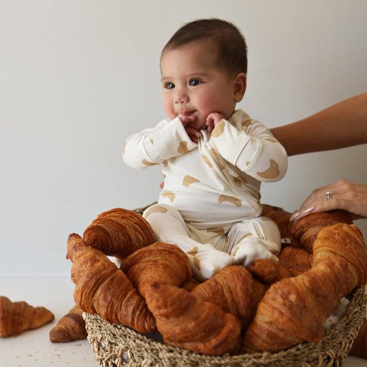 A baby in forever french baby Bamboo Zip Pajamas | Croissants sits in a basket of croissants, gently supported by an adult's hand against a plain background.