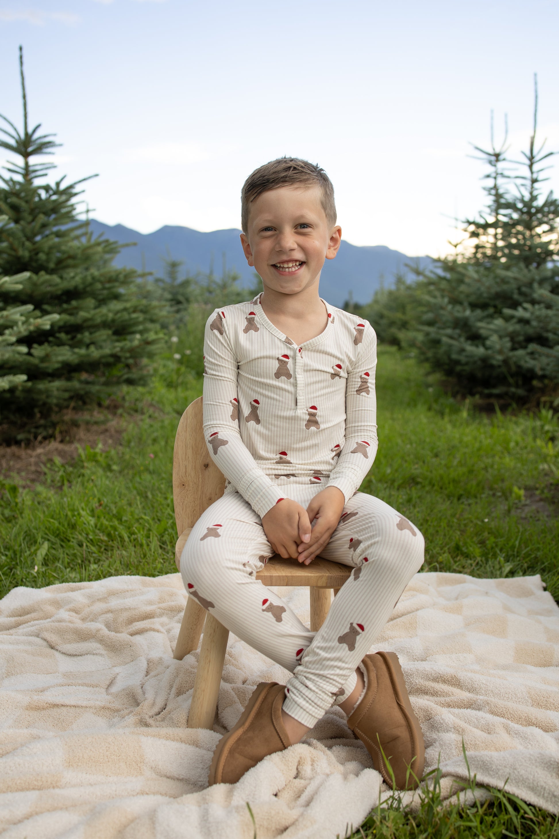A young boy wearing forever french baby's Bamboo Two Piece Pajamas in the Santa Teddy print sits on a wooden chair, smiling on a blanket outdoors among evergreen trees, with mountains and a blue sky in the background.