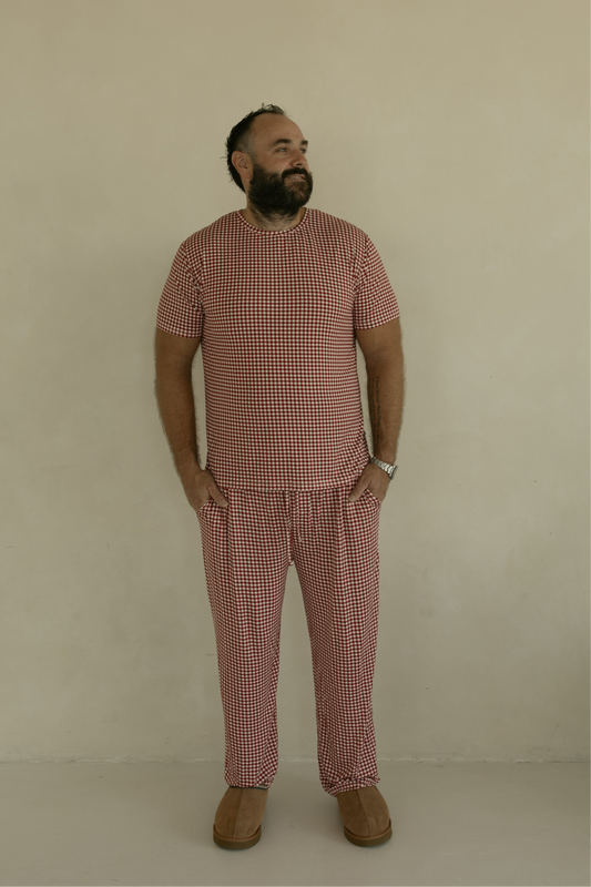 A bearded man stands against a plain light wall, wearing forever french baby's Men's Short Sleeve Bamboo Pajamas in Red Gingham, with hands in his pockets and brown slip-on shoes.