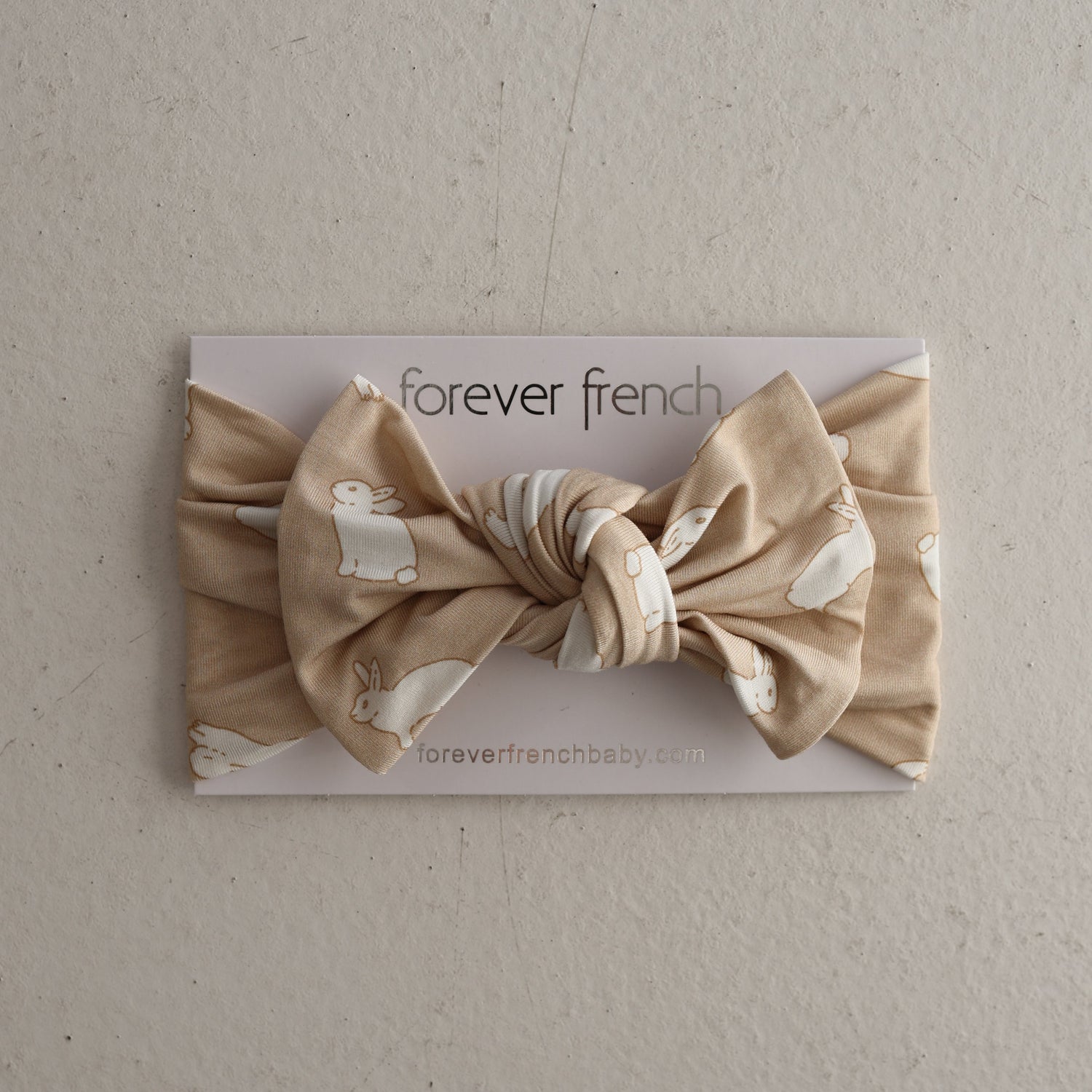 The Bamboo Head Wrap | Bunnies by forever french baby is a beige knotted baby bow headband featuring white bunny prints, shown on a “forever french baby” card against a neutral backdrop.