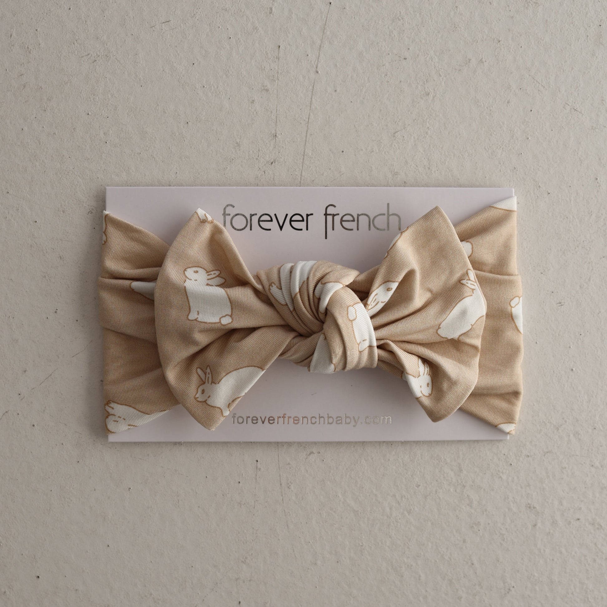 The Bamboo Head Wrap | Bunnies by forever french baby is a beige knotted baby bow headband featuring white bunny prints, shown on a “forever french baby” card against a neutral backdrop.