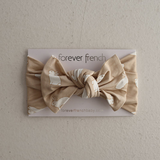 The Bamboo Head Wrap | Bunnies by forever french baby is a beige knotted baby bow headband featuring white bunny prints, shown on a “forever french baby” card against a neutral backdrop.