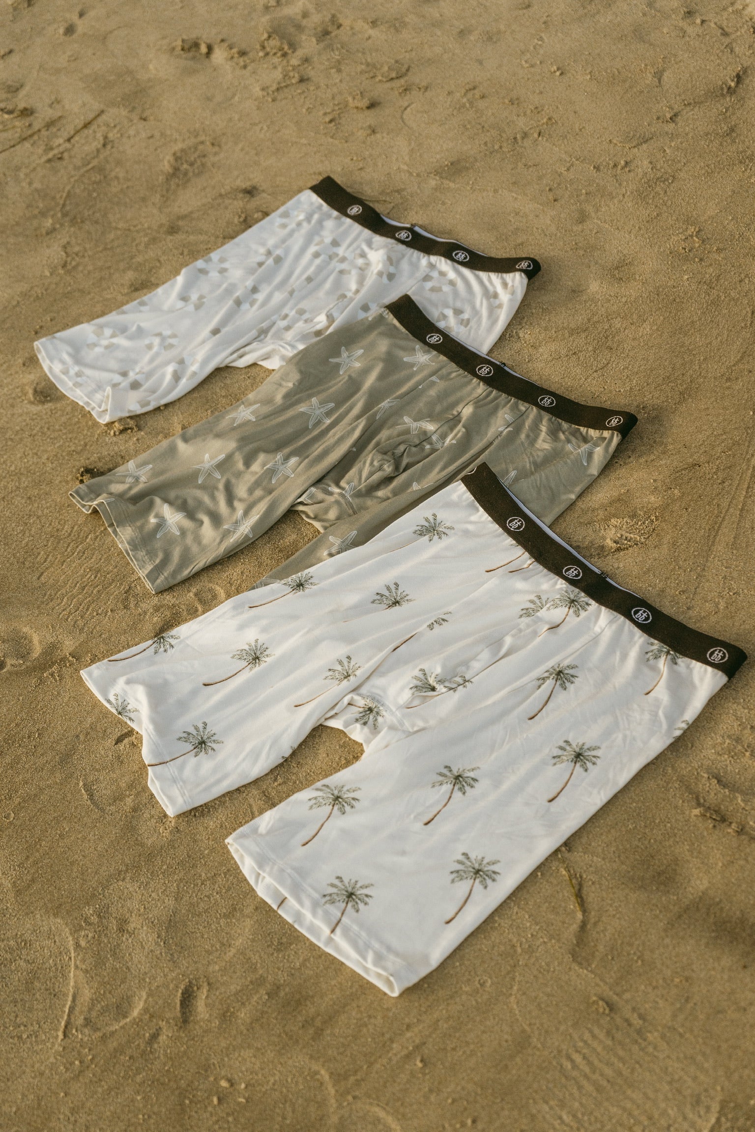Three pairs of forever french baby Men's Bamboo Boxers (3 Pack) in Lifesaver, Starfish & Palm Tree prints feature black waistbands and tropical designs, with two white and one olive green laid out on sandy ground.