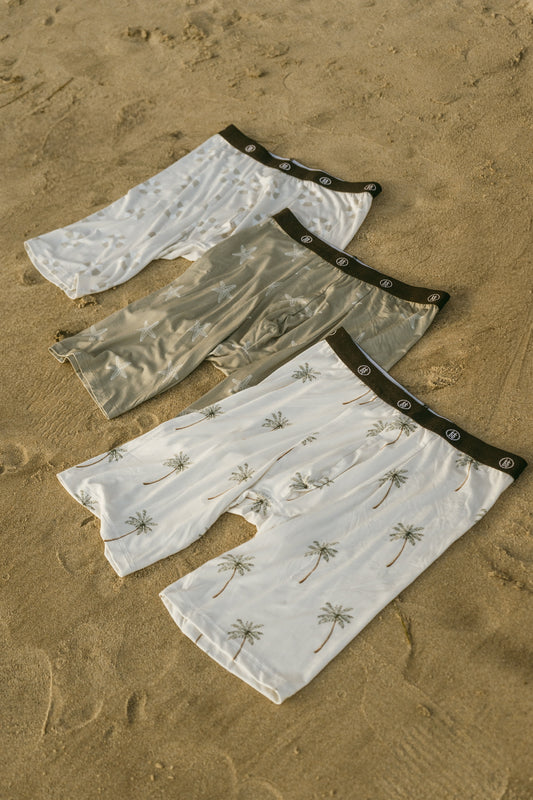 Three pairs of forever french baby Men's Bamboo Boxers (3 Pack) in Lifesaver, Starfish & Palm Tree prints feature black waistbands and tropical designs, with two white and one olive green laid out on sandy ground.