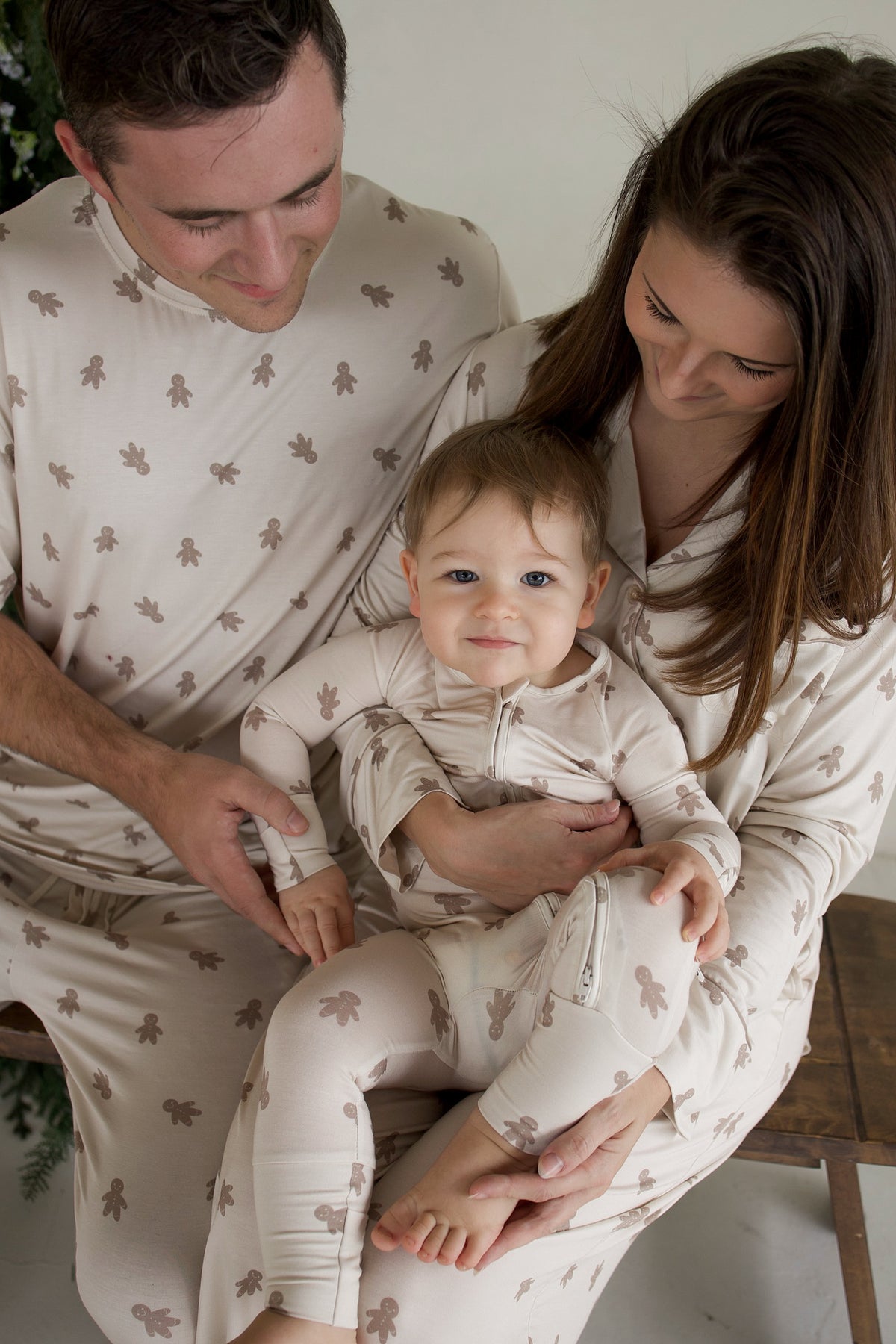 A man and woman holding a baby, all wearing matching beige Bamboo Zip Pajamas | Gingersnap featuring a repeating gingerbread man pattern.