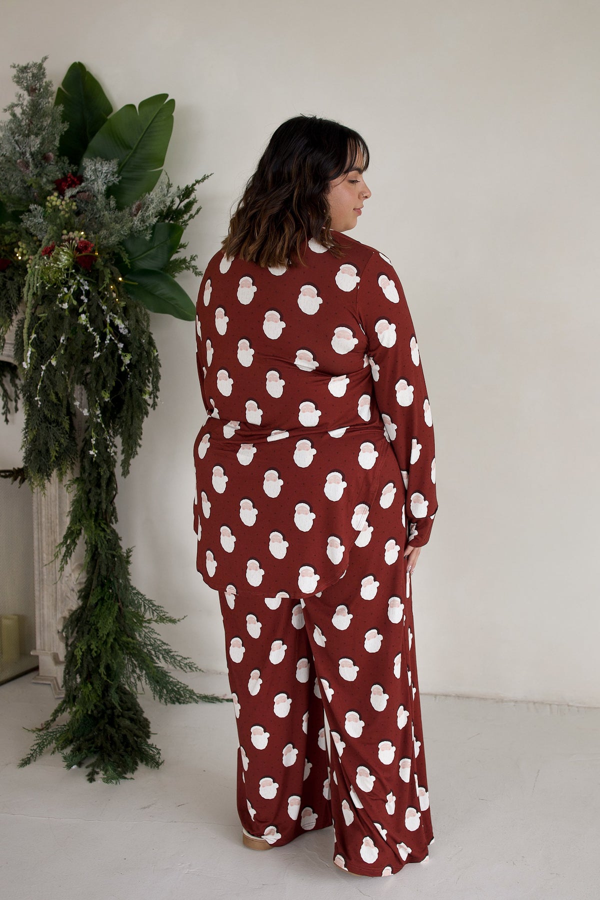 A person wearing a deep red Women’s Bamboo Button Up Lounge Set | Vintage Santa, featuring a long-sleeved button-up top and matching wide-leg pants with a repeating white Santa Claus print.