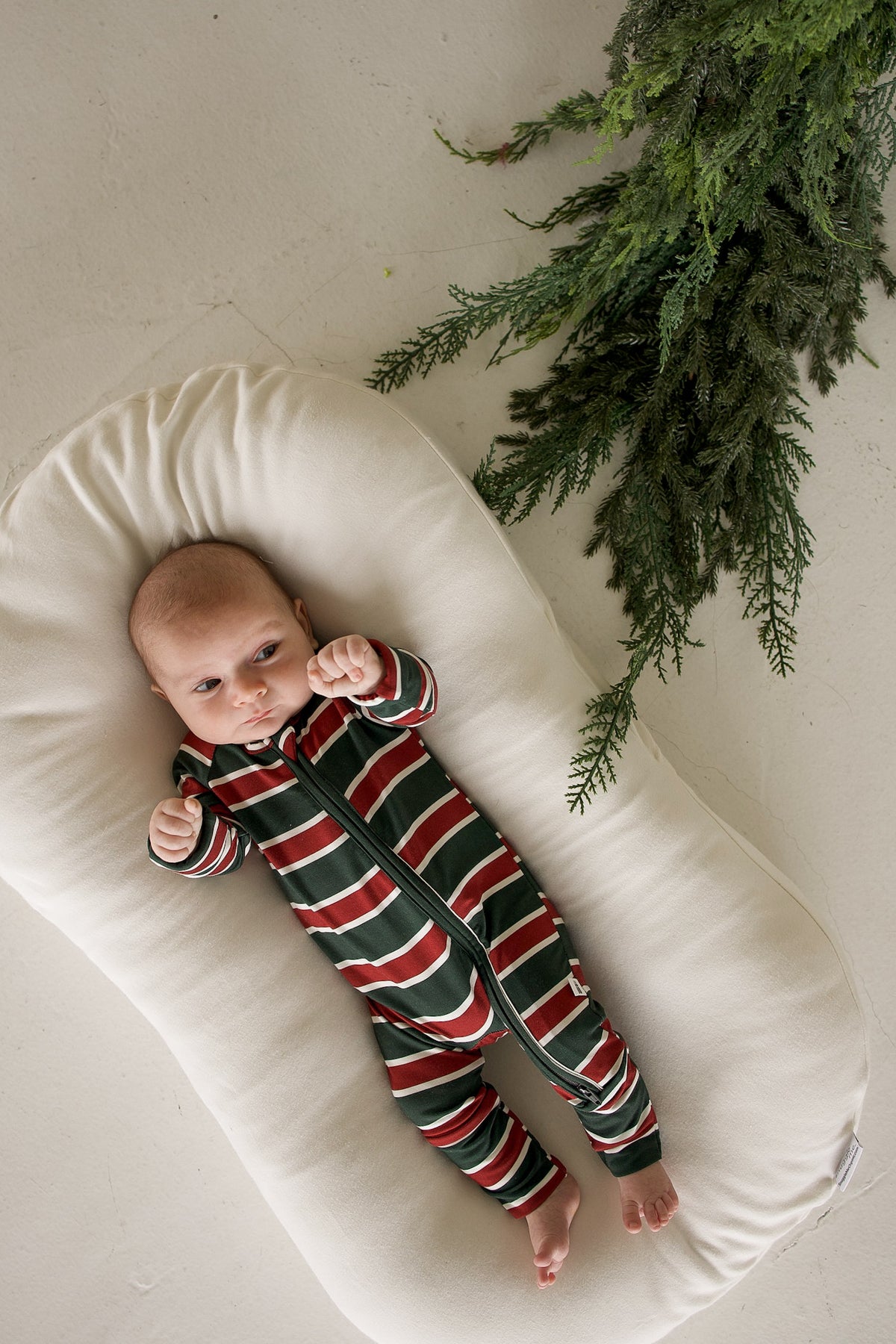 A baby wearing Bamboo Zip Pajamas | Holiday Stripe lies on a white lounger in the zippered sleeper.