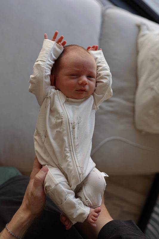 A baby in forever french baby's Bamboo Zip Pajamas | Dandelion is held upright with both arms raised and eyes half-closed, sitting on a light-colored couch.