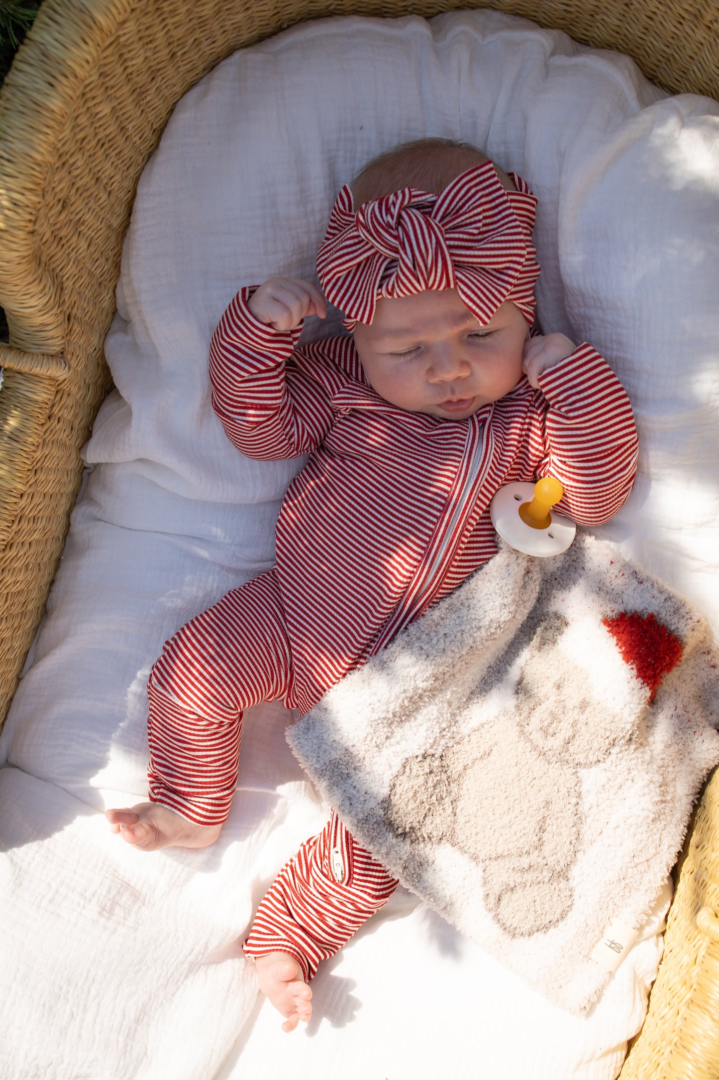 A baby in a basket wears a red-and-white striped outfit and matching headband, lying on a white blanket with a pacifier and the forever french baby Lovey | Santa Teddy, featuring an adorable Santa Teddy bear design.