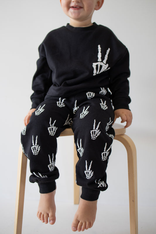 A young child sits barefoot on a light wooden stool against a plain white background, wearing the forever french baby Child Sweat Set | Midnight Skelly—a black children’s sweat set featuring playful white skeleton hand designs, perfect for everyday adventures.