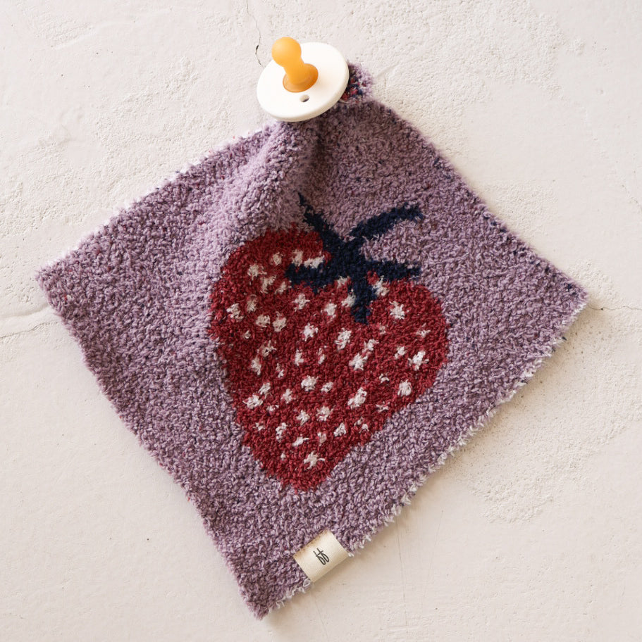 The forever french baby Lovey | Matilda features a purple, textured fabric with a large red strawberry and white dots, attached to a yellow pacifier and displayed on a light, rough surface.