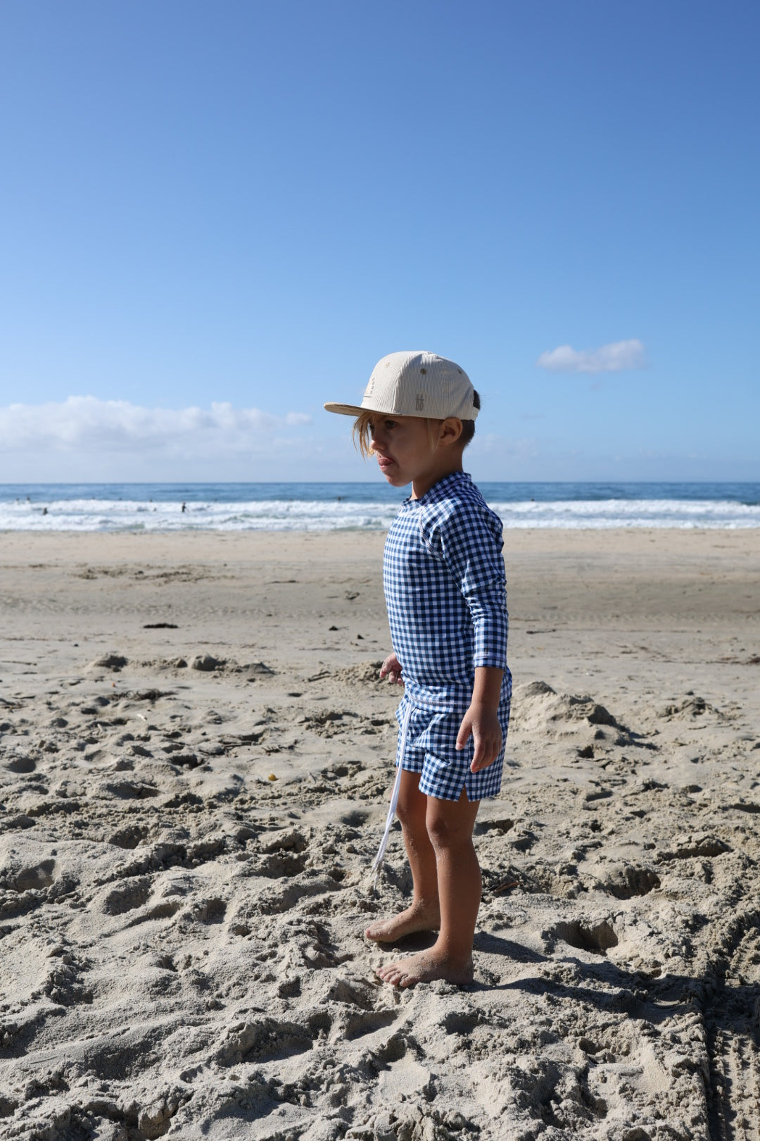 A young child, barefoot on the sand, gazes at the ocean wearing forever french baby's Child Board Short in Blue Gingham and a beige cap under a clear blue sky.