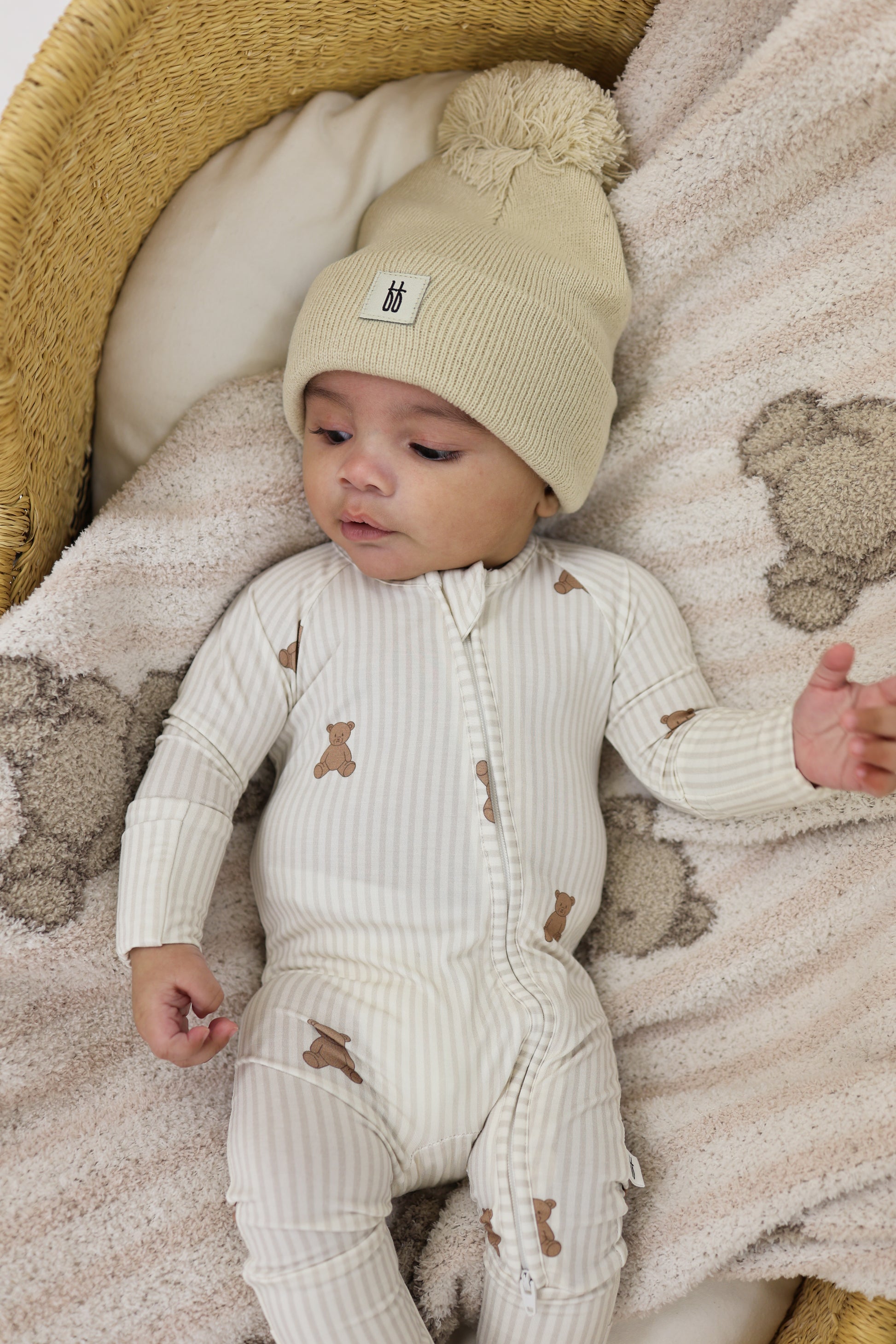 A baby wearing forever french baby's Bamboo Zip Pajamas | Teddy lies on a soft bear-face blanket in a wicker basket, topped with a cream knit hat.