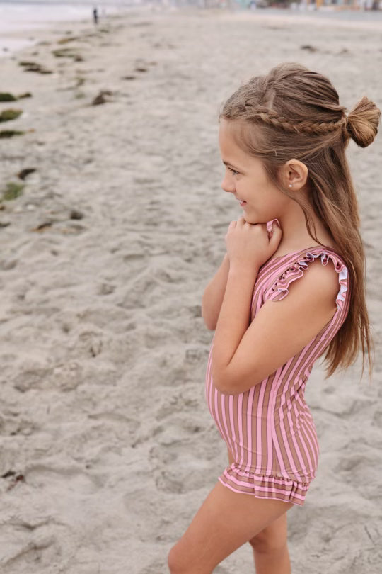 A young girl with long hair, styled in braids and a bun, is standing on a sandy beach. She is wearing the One Piece Girls Swim Suit | Boardwalk by forever french baby, featuring pink stripes and ruffled sleeves, and is smiling while looking towards the ocean. The beach extends into the distance with scattered rocks and seaweed.