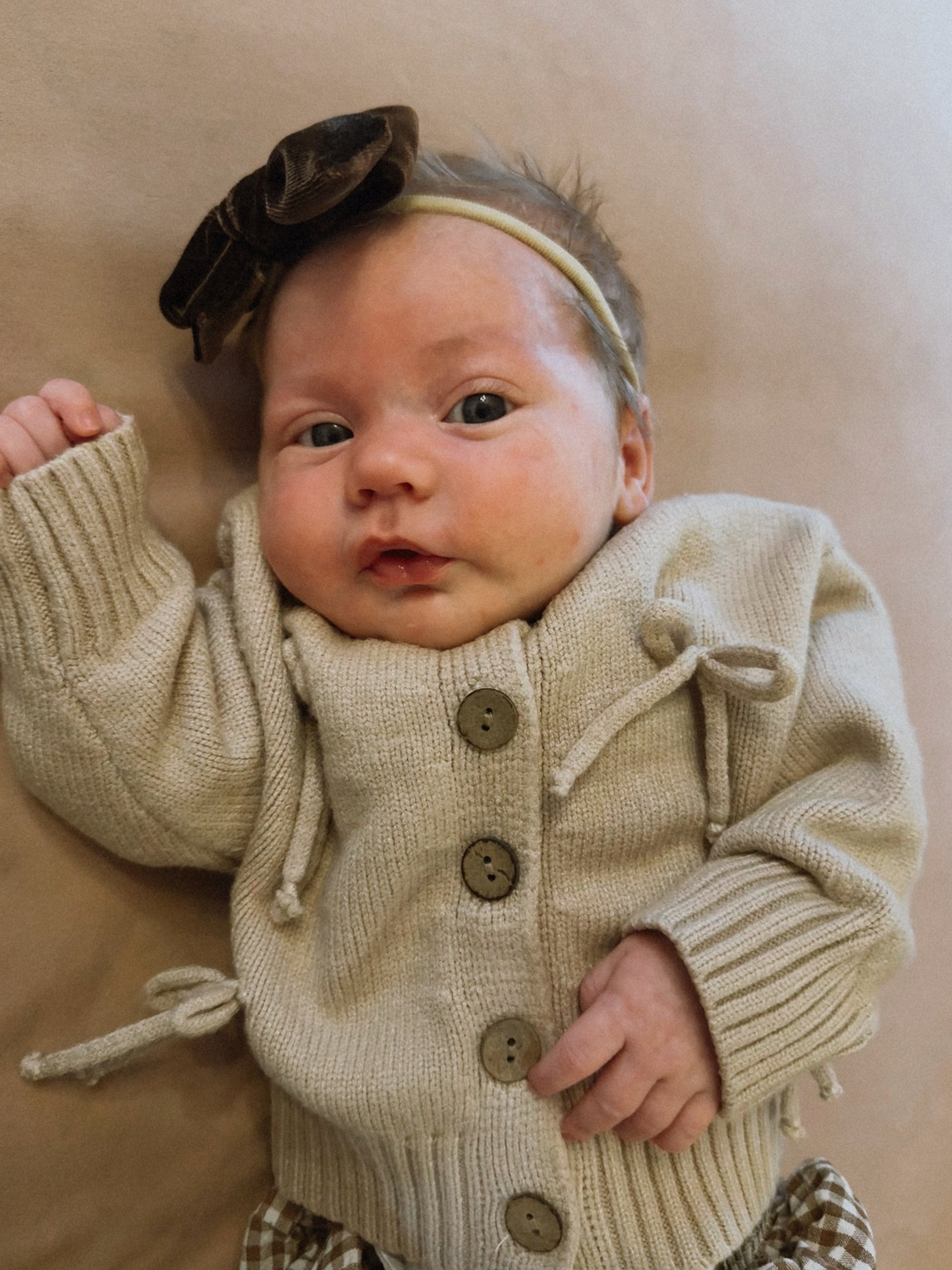 A baby in a forever french baby Knit Cardigan | Bows—beige, with large buttons and bows—paired with a brown bow headband, lies on a soft beige surface, gazing up with one arm raised.