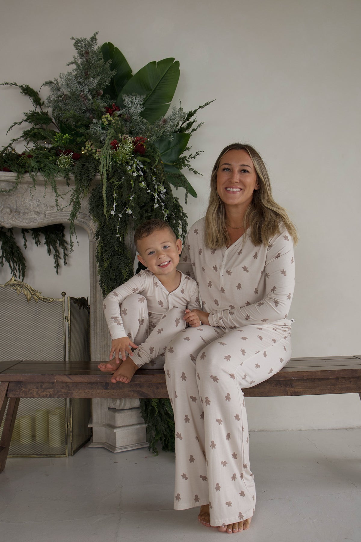 A woman and a young boy sit together on a wooden bench, wearing matching beige Bamboo Two Piece Pajamas | Gingersnap by forever french baby, featuring a brown gingerbread man pattern.