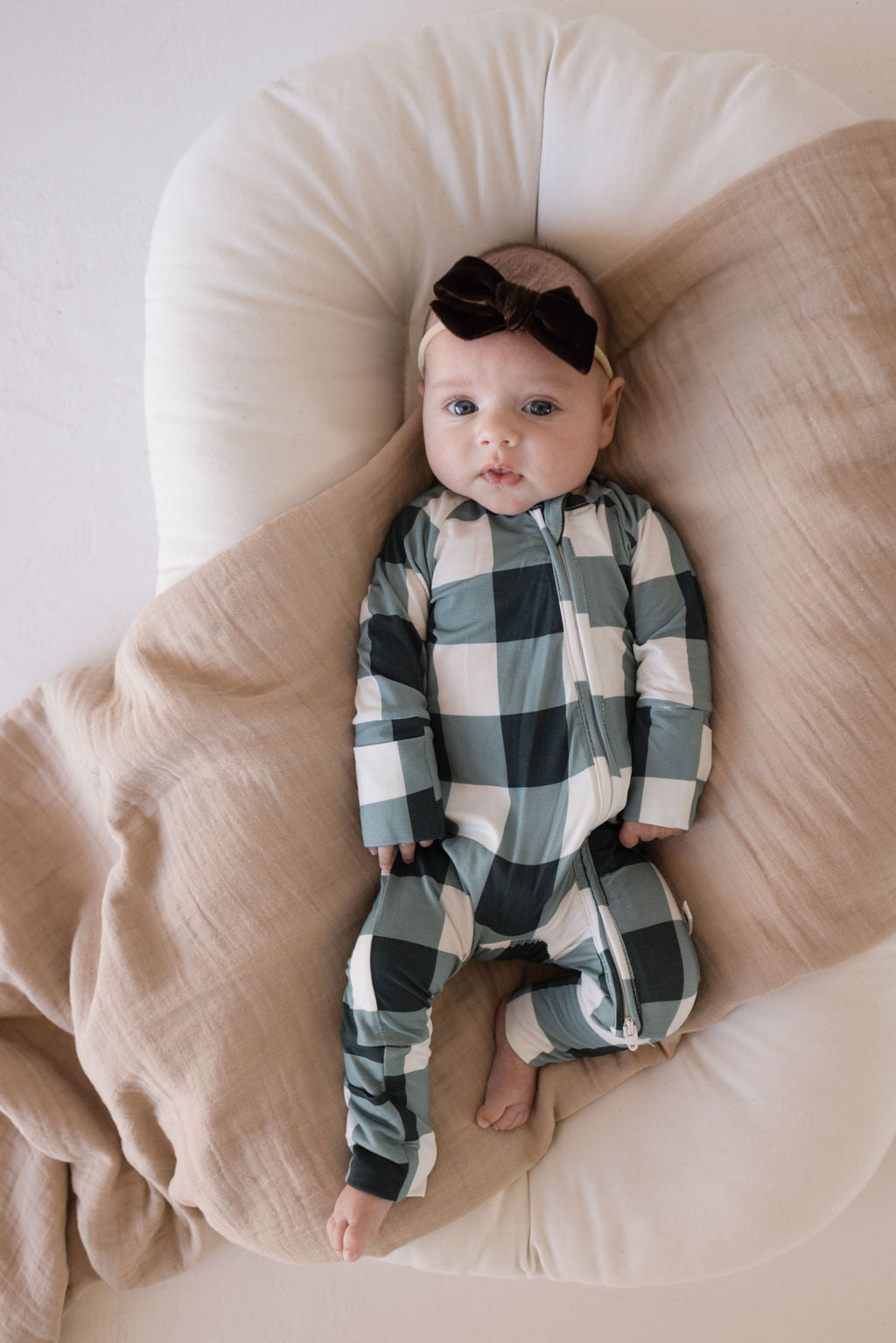 A baby is lying on a cushioned surface, partially covered by a beige blanket, wearing the Bamboo Zip Pajamas in Evergreen Gingham from forever french baby, along with a black bow headband. The onesie is made of breathable fabric and features green and white checkered patterns. The baby has a calm expression while looking up.