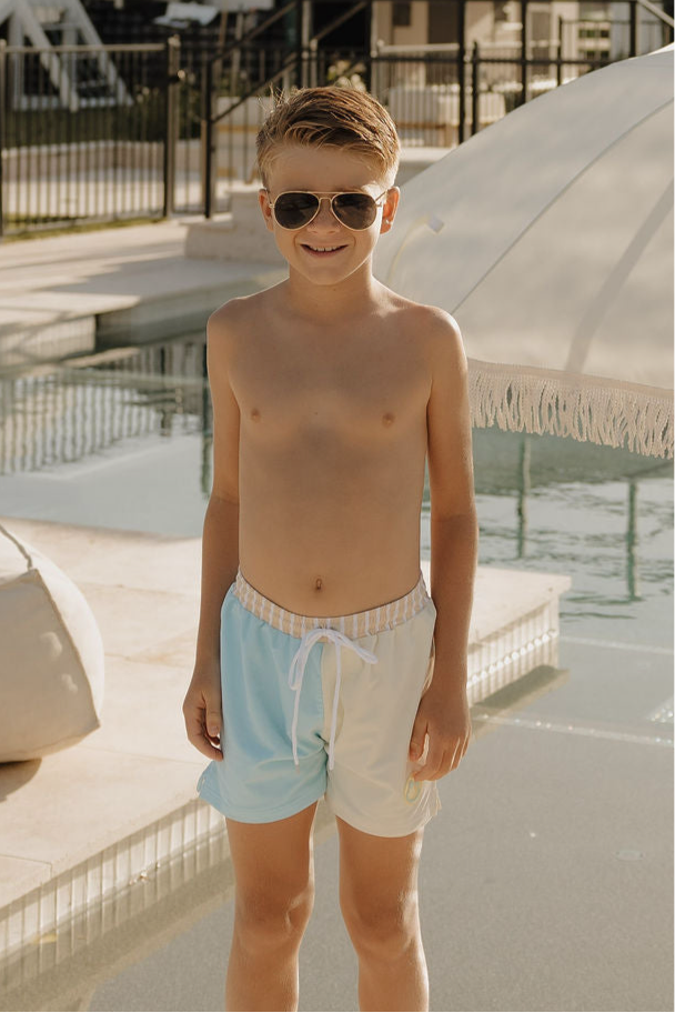 A young boy in lolo webb 2’s Child Board Short | Surf & Sand and sunglasses smiles by the pool, with a large white umbrella and poolside furniture in the background.