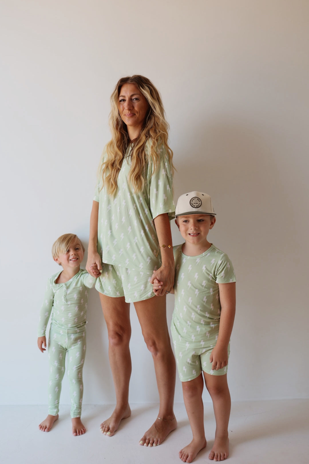 A woman and two young children stand barefoot against a plain background, all wearing matching Bamboo Short Set | Flash pajamas by forever french baby, featuring light green fabric with a white animal print. The woman holds one child’s hand as they smile.