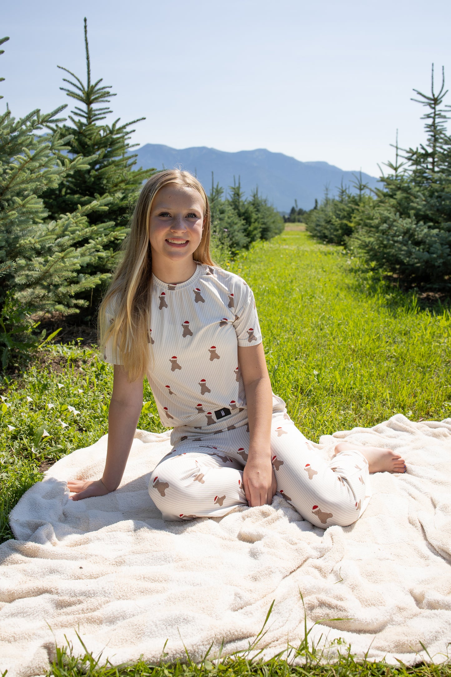 A girl wearing the Child Boxy Tee Lounge Set | Santa Teddy by forever french baby, made from hypo-allergenic bamboo fabric, sits on a blanket in a grassy field among pine trees, with mountains visible in the background under a clear sky.
